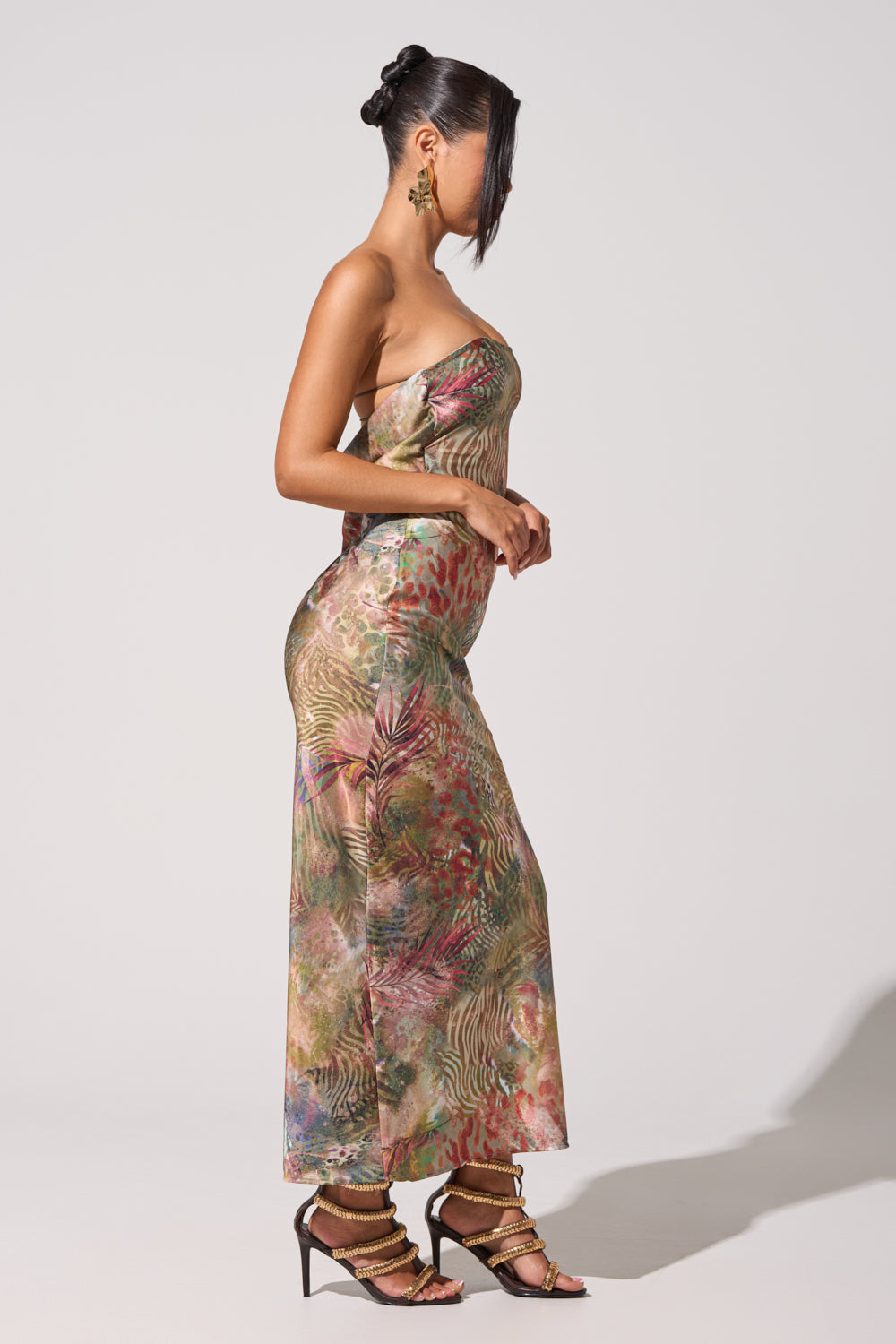 A woman with an updo stands in profile against a plain light backdrop, wearing the IT GIRL SATIN MAXI DRESS with a vibrant tropical print and black high-heeled sandals. 4