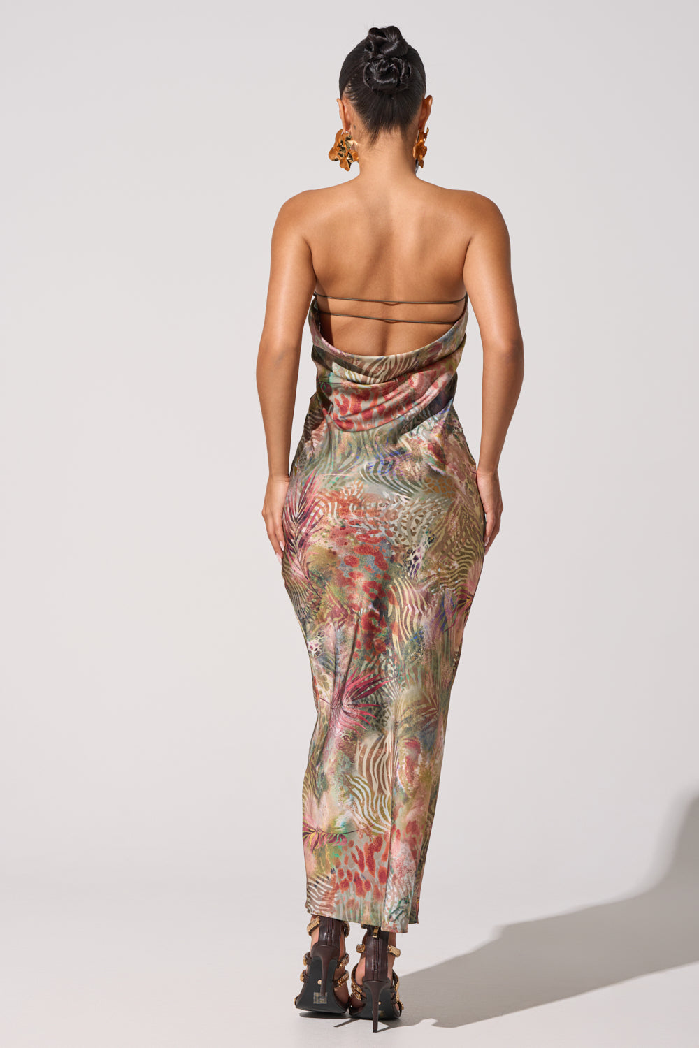 A person faces away, wearing the IT GIRL SATIN MAXI DRESS with a bold tropical print and cowl back, paired with black high-heeled sandals. Their hair is in a neat bun and they wear statement earrings, set against a plain light background. 5