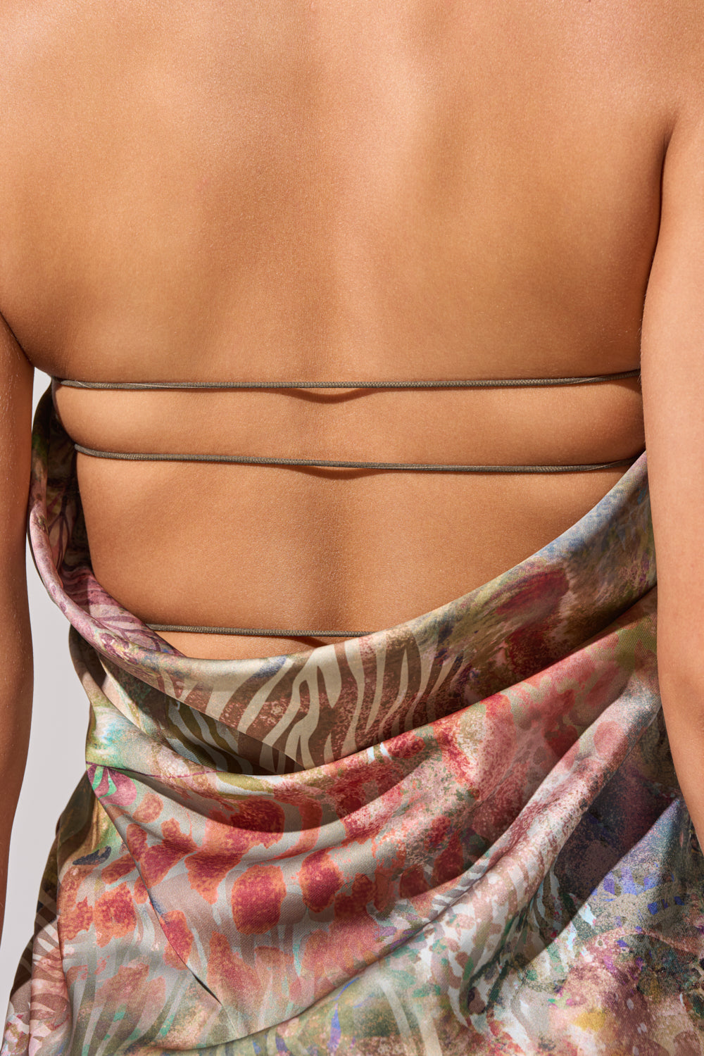 A close-up of a person’s bare upper back showcases the IT GIRL SATIN MAXI DRESS, featuring thin horizontal straps and a colorful tropical print with floral and abstract red, green, and beige designs. 7