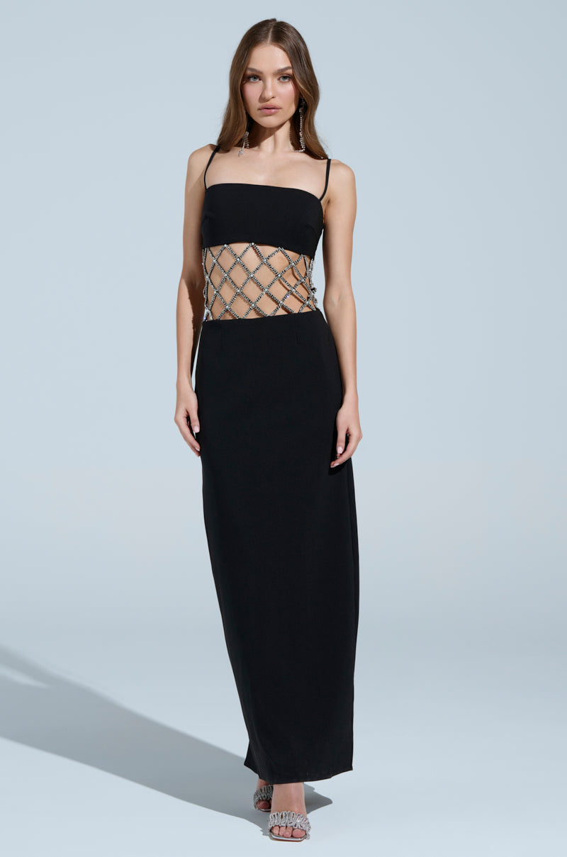 A person models the TAKE IT BLACK MIDI DRESS WITH RHINESTONE CUTOUT, featuring spaghetti straps and a detailed crisscrossed rhinestone pattern at the midsection, set against a plain soft blue background.