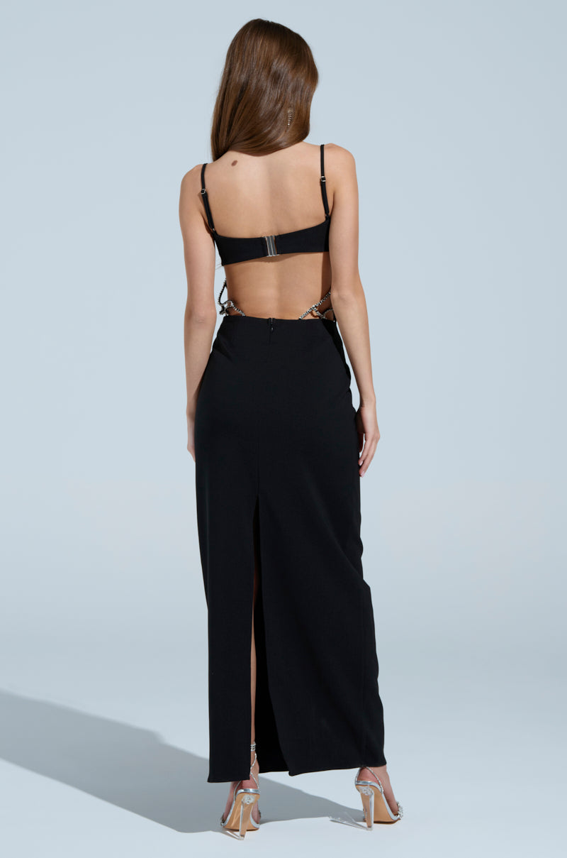 A long-haired woman showcases the TAKE IT BLACK MIDI DRESS WITH RHINESTONE CUTOUT. The curve-hugging, sleeveless design features a rhinestone cut-out back and sleek slit, paired with silver high heels against a neutral background.