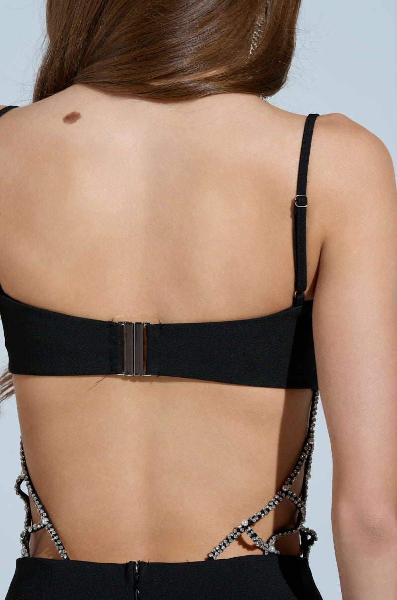 A person with long brown hair is seen from the back in the TAKE IT BLACK MIDI DRESS WITH RHINESTONE CUTOUT, showcasing rhinestone details on the open sides and straps. The figure-flattering silhouette highlights their contours, while a noticeable mole on their upper back adds intrigue.