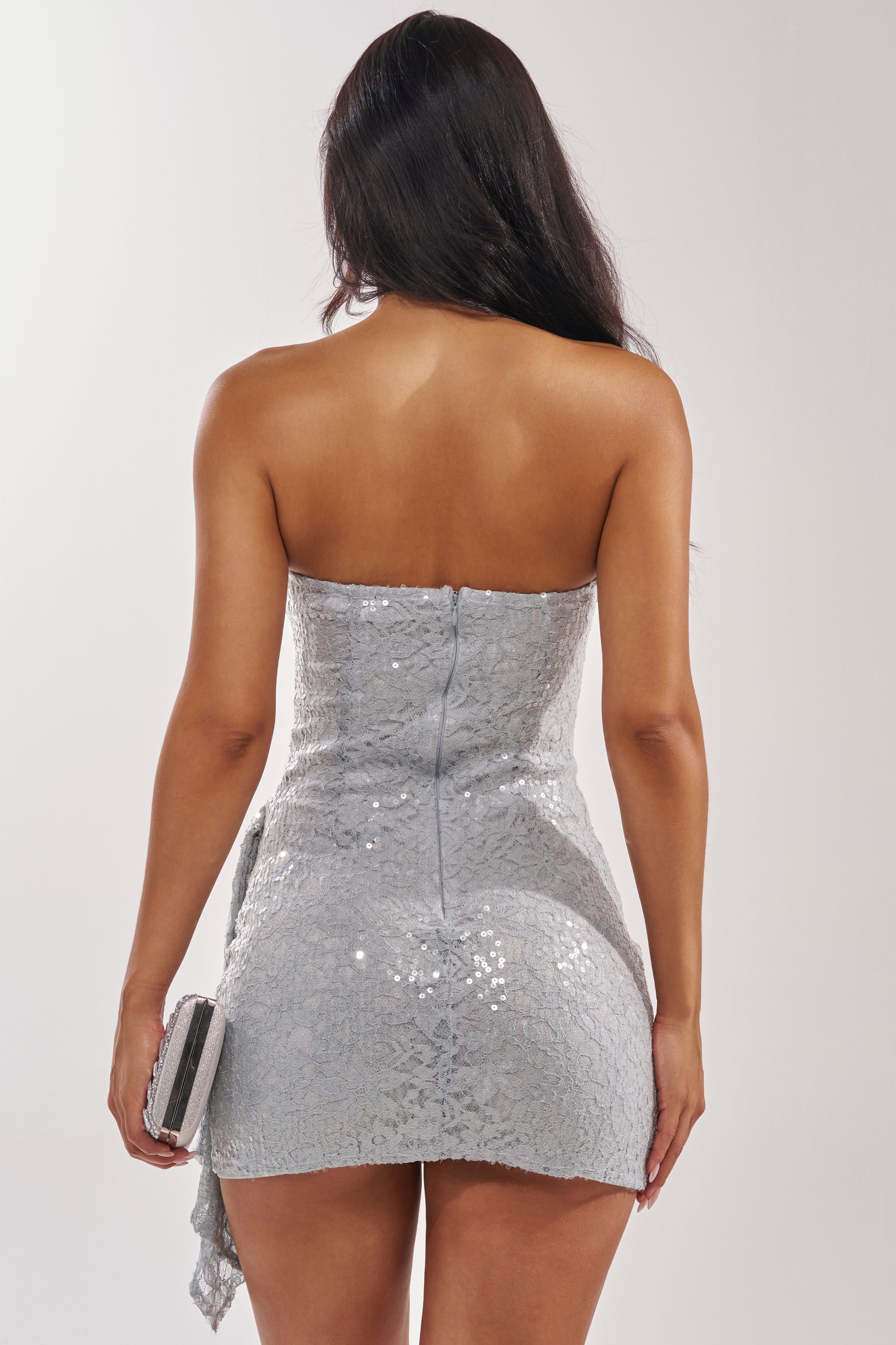 A woman with long dark hair faces away, wearing the LEAVE QUITE AN IMPRESSION LACE MINI DRESS IN LIGHT BLUE with a bodycon fit, holding a small silver clutch against a plain light background. 6