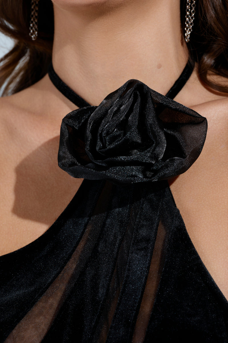 Close-up of a person wearing the SEE THROUGH ME MAXI DRESS, a black velvet maxi with a large fabric rose at the neckline. The dress has a sheer, textured mermaid-like appearance, paired with long dark hair and small hoop earrings.