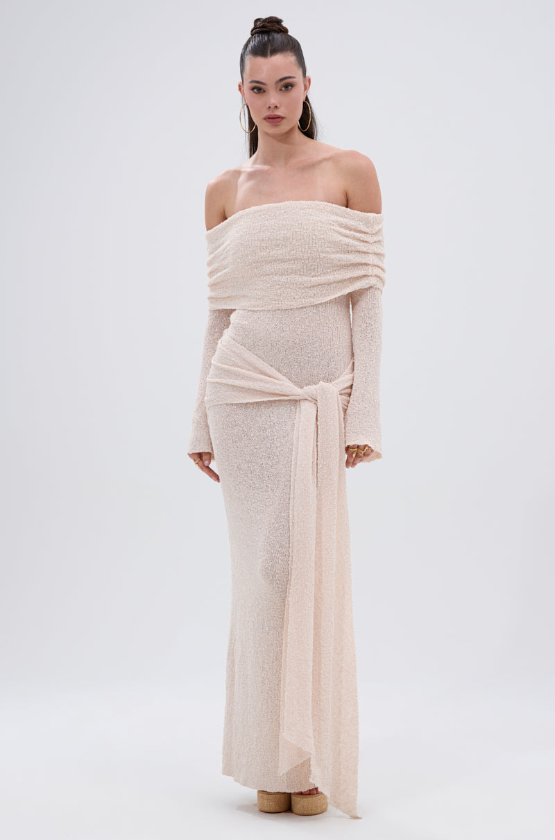 A woman models the IN YOUR DREAMS KNIT MAXI DRESS—an off-the-shoulder, long-sleeved maxi made from textured knit with a draped sash waist—standing against a simple light background.