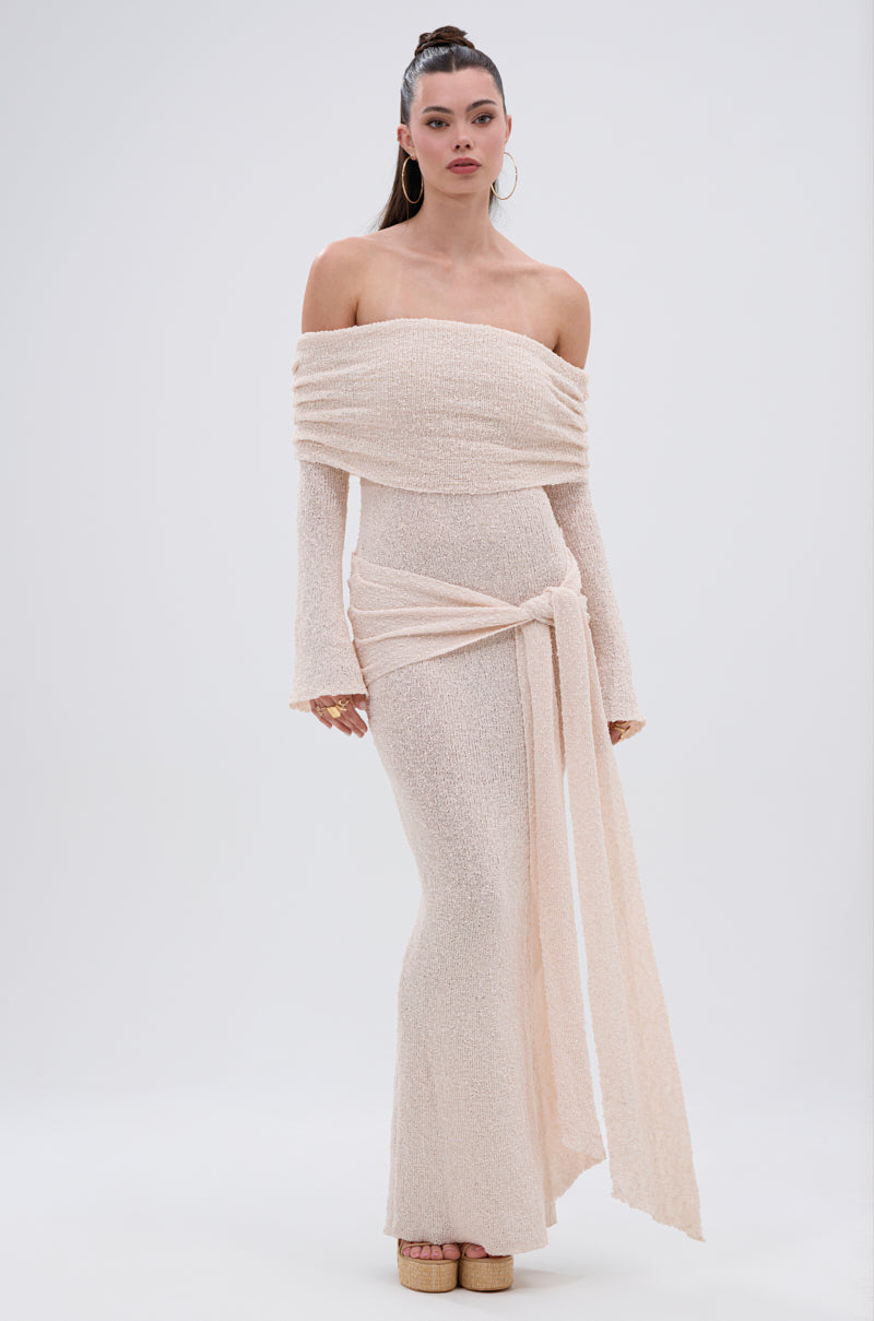 A woman stands against a plain light background wearing the IN YOUR DREAMS KNIT MAXI DRESS, an off-the-shoulder, long-sleeve dress with a wrapped waist in textured cream knit, paired with platform shoes and large hoop earrings.