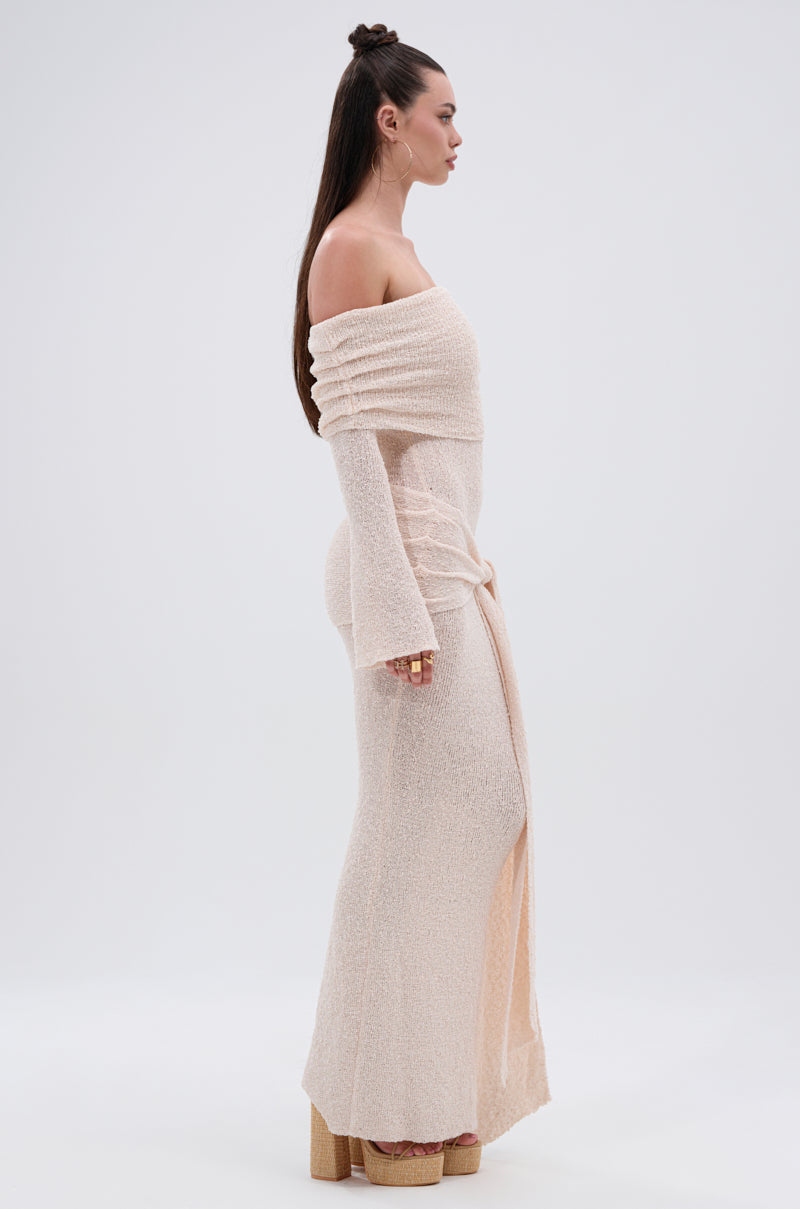 A woman stands in profile against a plain white background wearing the IN YOUR DREAMS KNIT MAXI DRESS—an off-the-shoulder, long-sleeve maxi with a waist wrap detail—paired with platform shoes. Her long, straight hair is styled half-up.