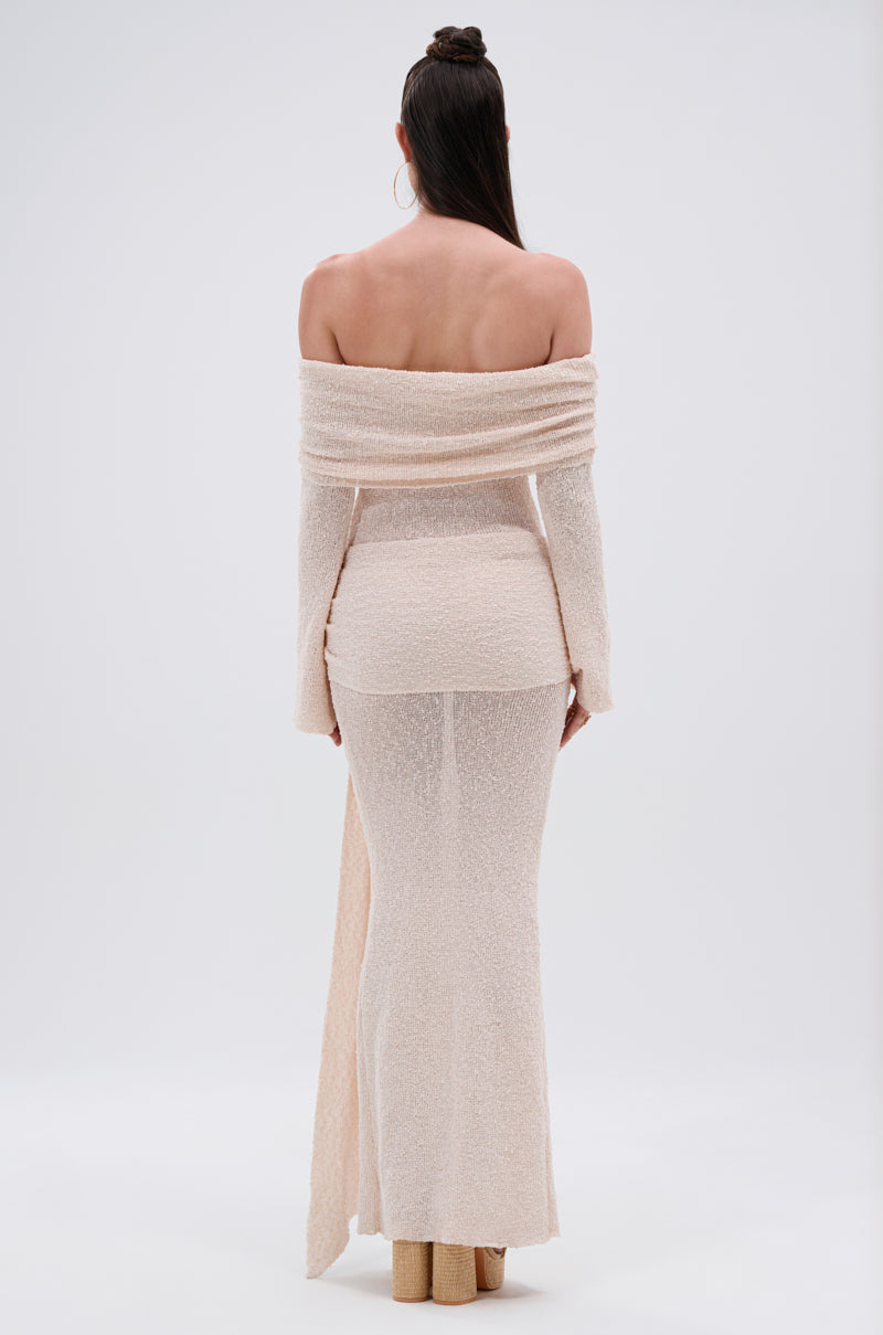 A woman with a sleek top knot faces away from the camera in the IN YOUR DREAMS KNIT MAXI DRESS, an off-the-shoulder, long-sleeve, textured beige maxi dress that reaches the floor. The background is plain white.