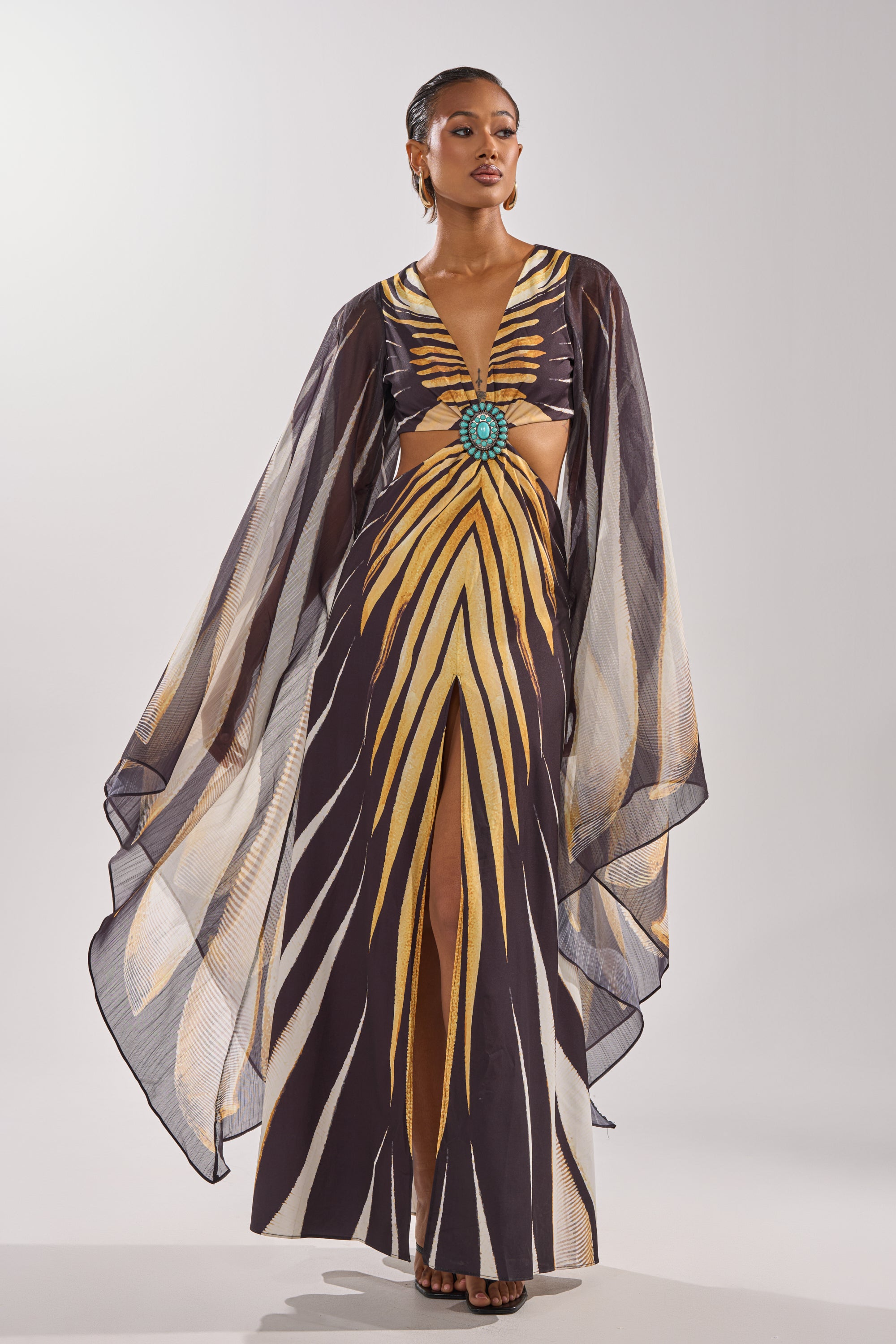A woman models the SHEER OPULENCE CUTOUT MAXI DRESS, a chiffon maxi featuring wide, sheer sleeves, bold yellow and brown stripes, a turquoise waist embellishment, front slit, drop earrings, and slicked-back hair.