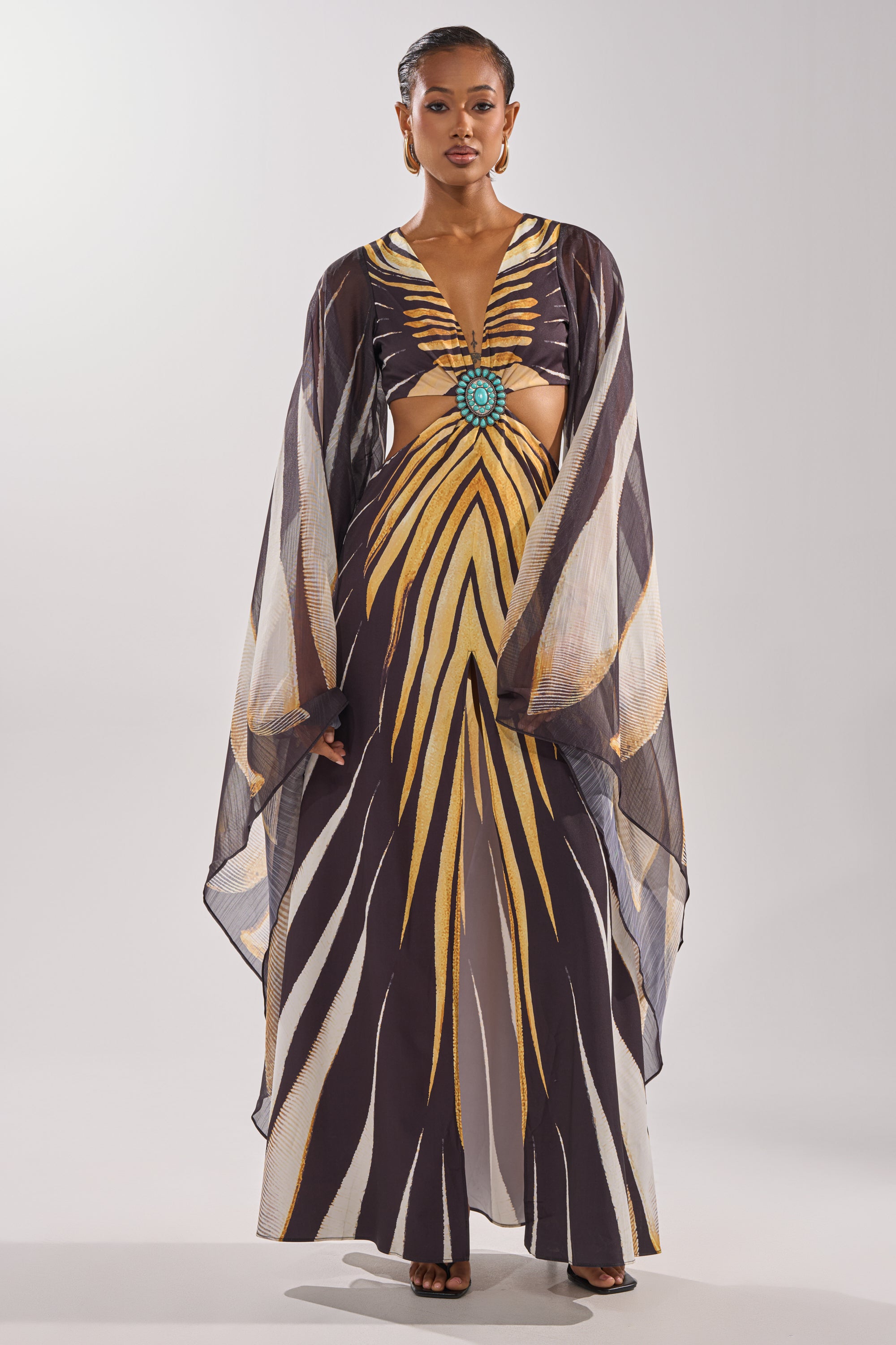 A woman wears the SHEER OPULENCE CUTOUT MAXI DRESS by AKIRA Label, featuring bold yellow and black geometric patterns, sheer flowing sleeves, a front cut-out, and a large turquoise brooch at the bodice.