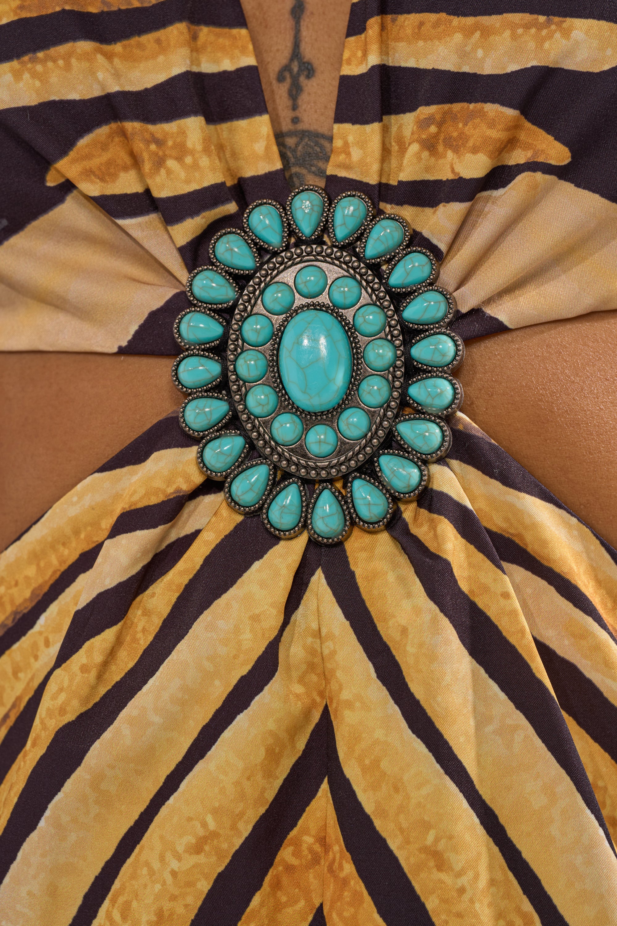 A close-up of the SHEER OPULENCE CUTOUT MAXI DRESS features a turquoise and silver brooch on yellow and brown striped chiffon, with a skin-baring cutout and a small tattoo visible above the brooch.