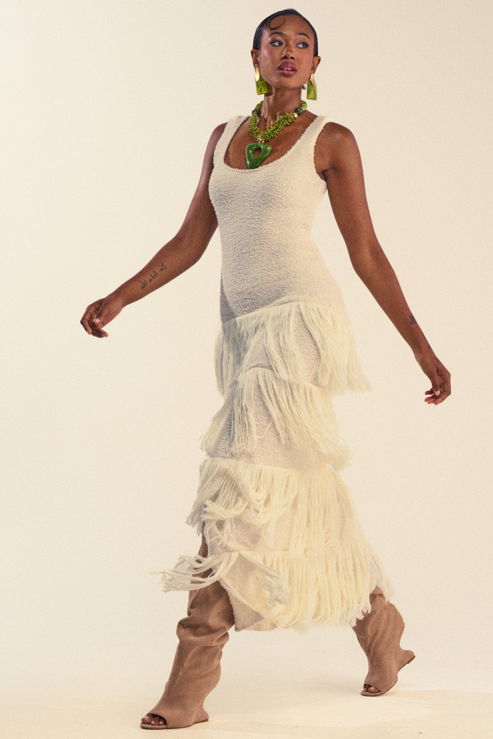 Wearing the ROGUE HEART KNIT FRINGE MAXI DRESS, a woman poses against a light background styled with beige slouchy boots and bold green jewelry. The cozy chic maxi dress features textured white fringe and tiered details for a striking look.