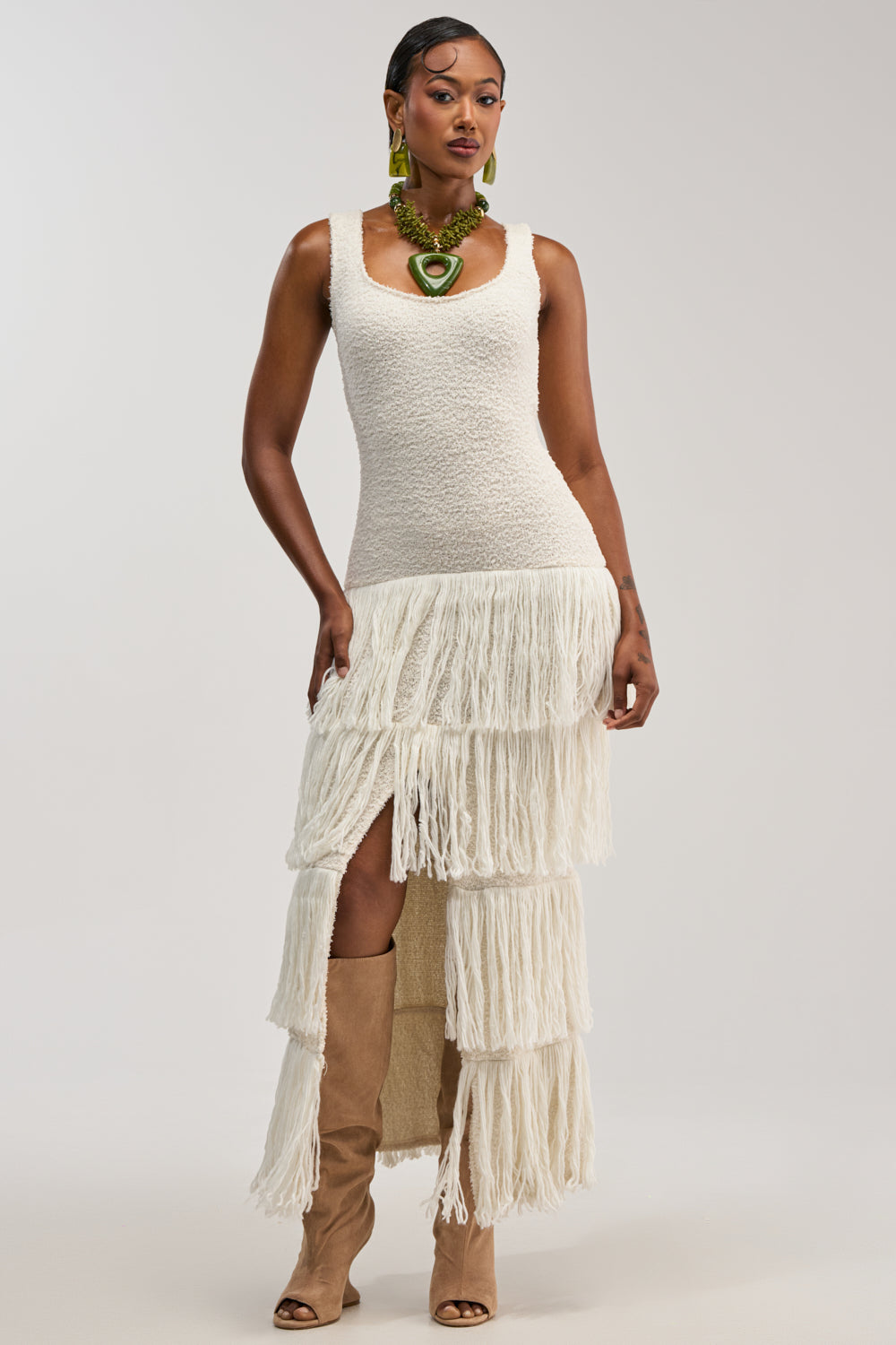 Wearing the ROGUE HEART KNIT FRINGE MAXI DRESS—a sleeveless white textured dress with layered fringe that's perfect for winter bridal events or a date night—styled with tall tan boots and bold green jewelry, she stands against a light background.