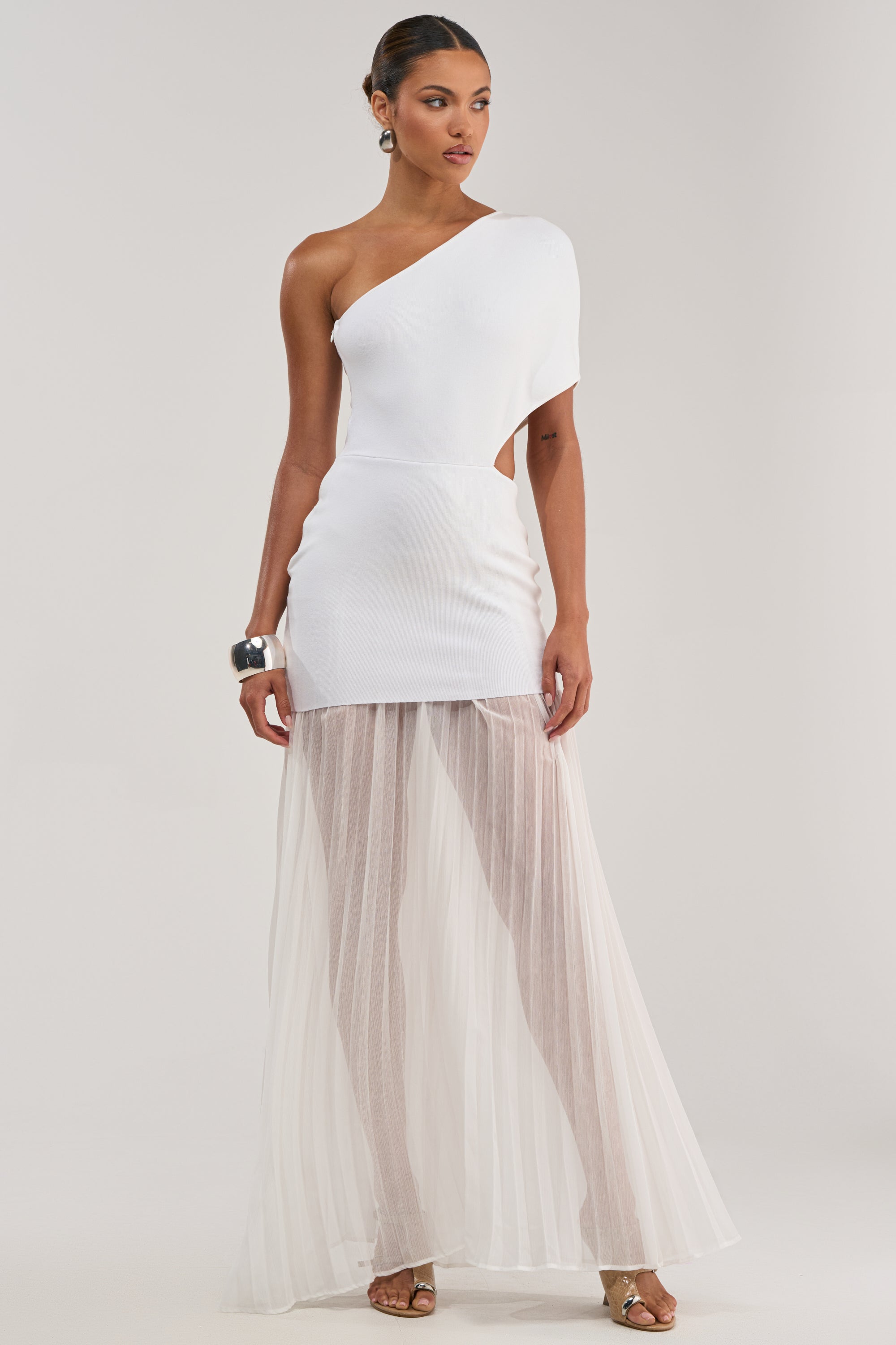 A woman poses in the FINAL BOW ASYMMETRICAL MAXI DRESS IN WHITE, a bridal-inspired one-shoulder style with a fitted mini skirt and sheer pleated maxi overlay, styled with silver earrings, a cuff bracelet, and strappy heels against a plain light background.