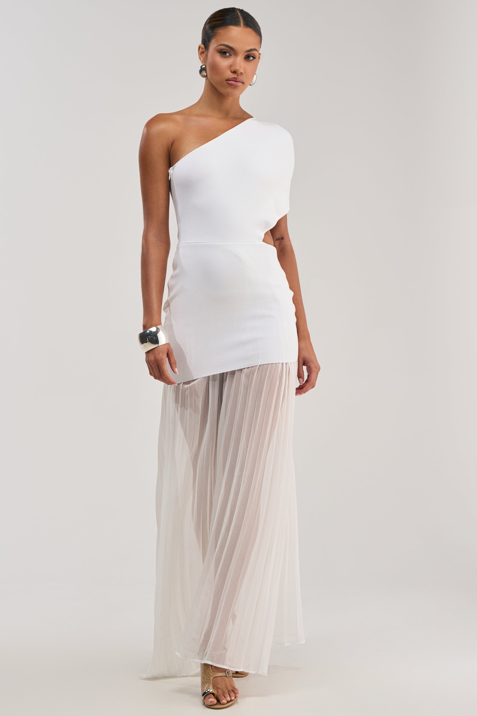 A woman models the FINAL BOW ASYMMETRICAL MAXI DRESS IN WHITE, featuring a chic asymmetrical neckline and sheer pleated skirt, styled with metallic sandals and a chunky bracelet against a plain background.