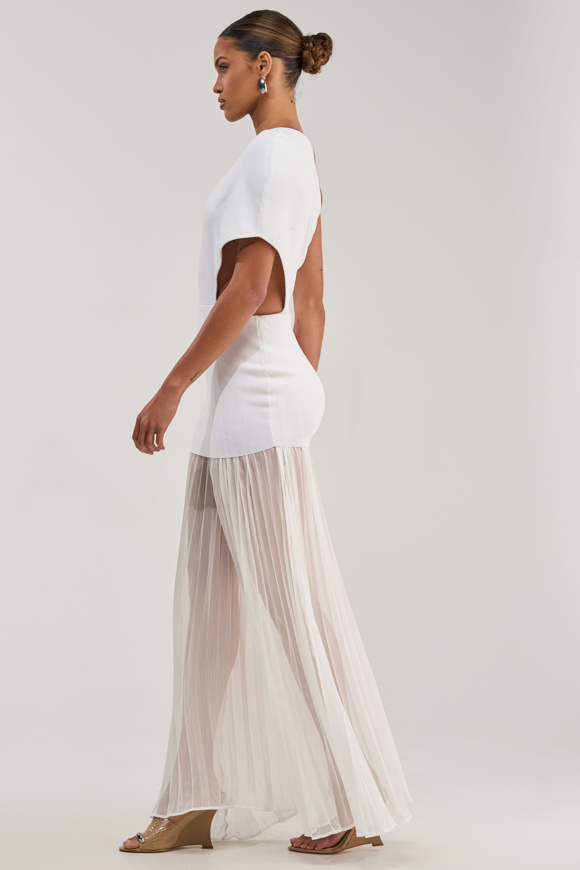 A woman in the FINAL BOW ASYMMETRICAL MAXI DRESS IN WHITE, featuring short sleeves, side cutouts, and a sheer pleated skirt, stands in profile against a plain background with tan wedge sandals and her hair in a neat bun.