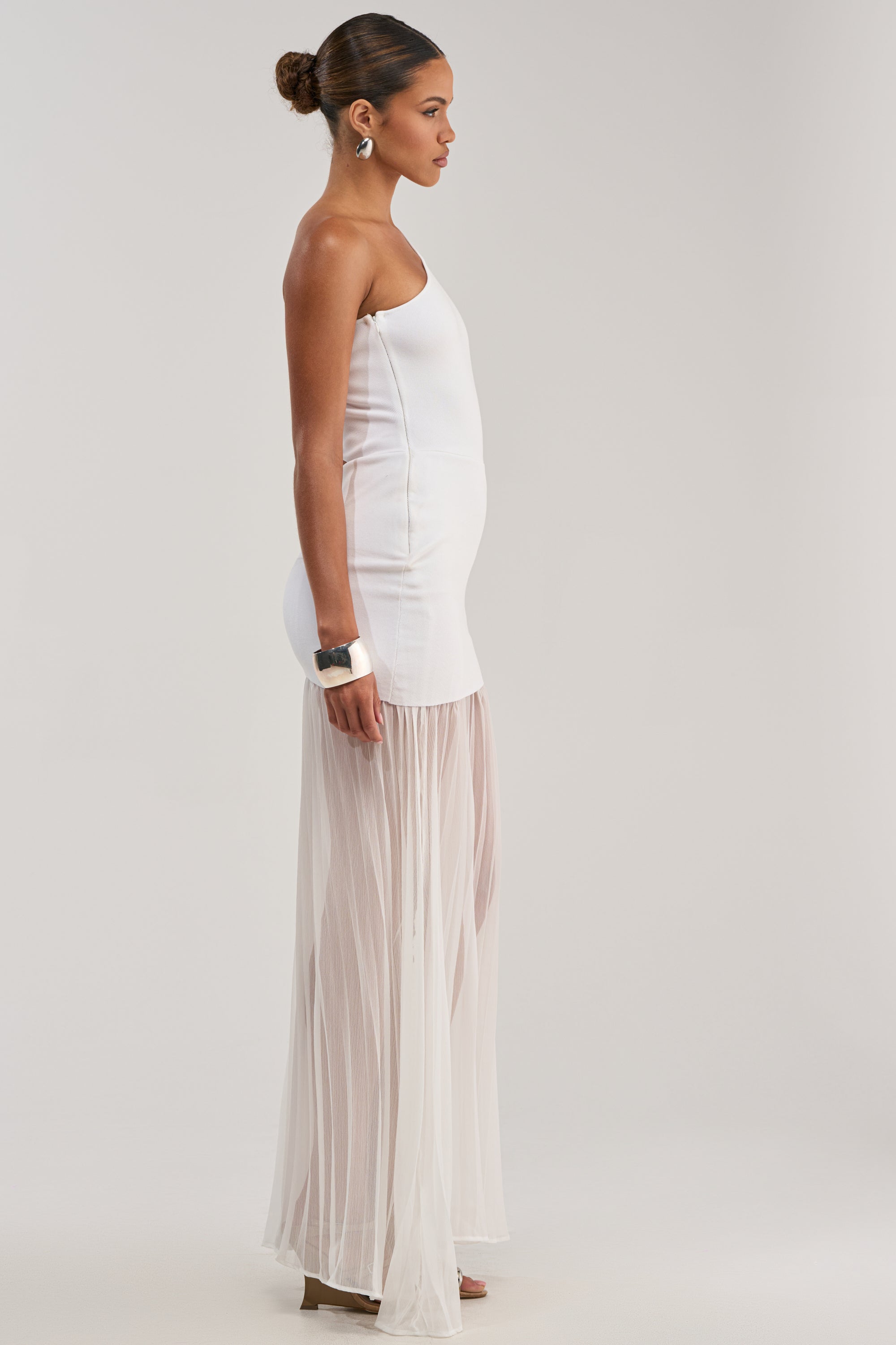 A woman stands in profile wearing the FINAL BOW ASYMMETRICAL MAXI DRESS IN WHITE, featuring an asymmetrical neckline, fitted bodice, and sheer flowing skirt. Her hair is in a neat bun with hoop earrings and a chunky bracelet for an elegant look.