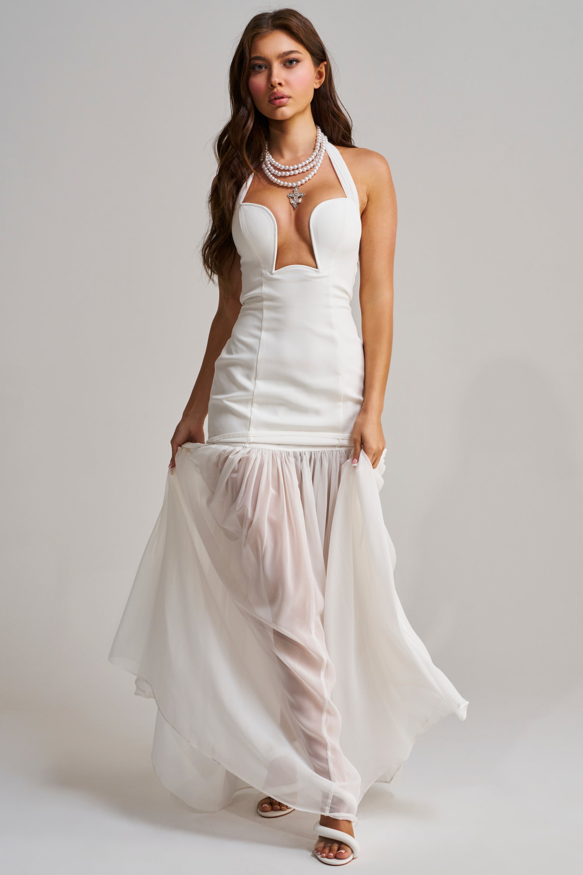 A woman wears the ANGEL ENERGY MAXI DRESS IN WHITE featuring a halter neck, deep neckline, fitted bodice, and sheer flowing skirt. She has long wavy hair, layered necklaces, and stands against a plain light background.