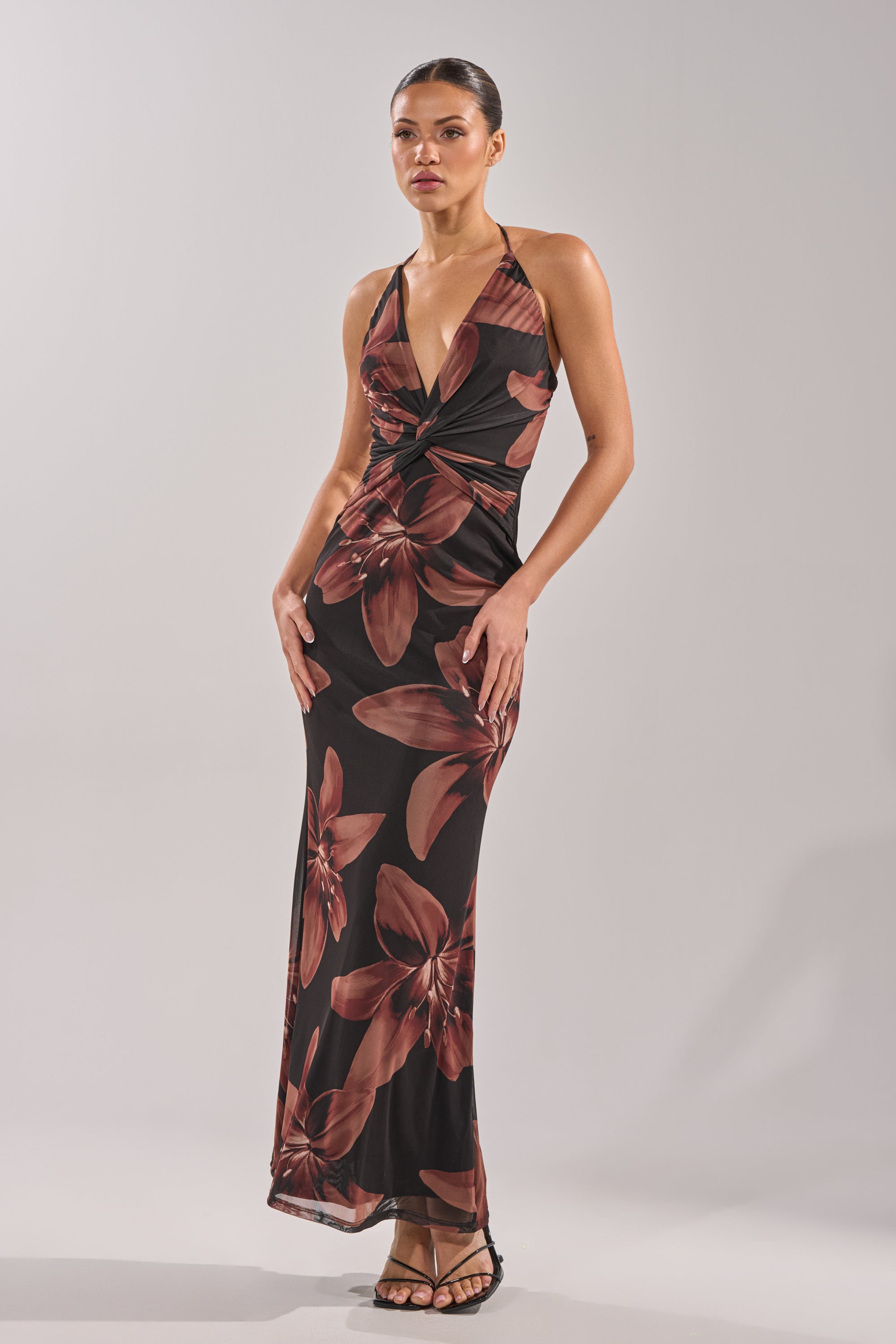 A woman stands confidently in strappy heels, wearing the FLOWER CHILD MAXI DRESS— a sleeveless, floor-length floral print with large red blooms, halter neckline, and open-back detail—against a plain light background. 2