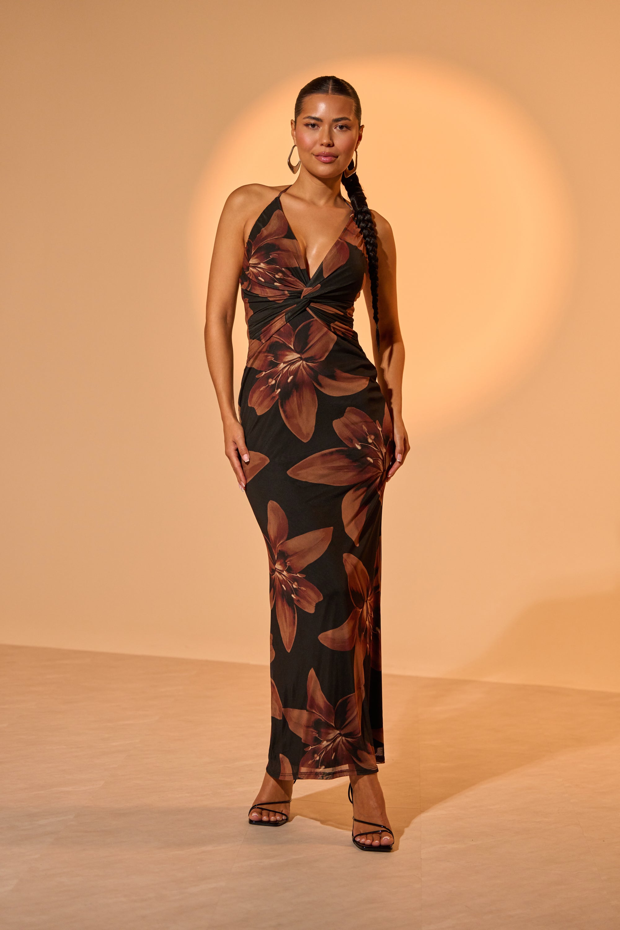 A woman stands confidently in the FLOWER CHILD MAXI DRESS, a halter-neck maxi with large brown floral prints, styled with hoop earrings and black heels. Her long braid falls over one shoulder against a beige background. 0