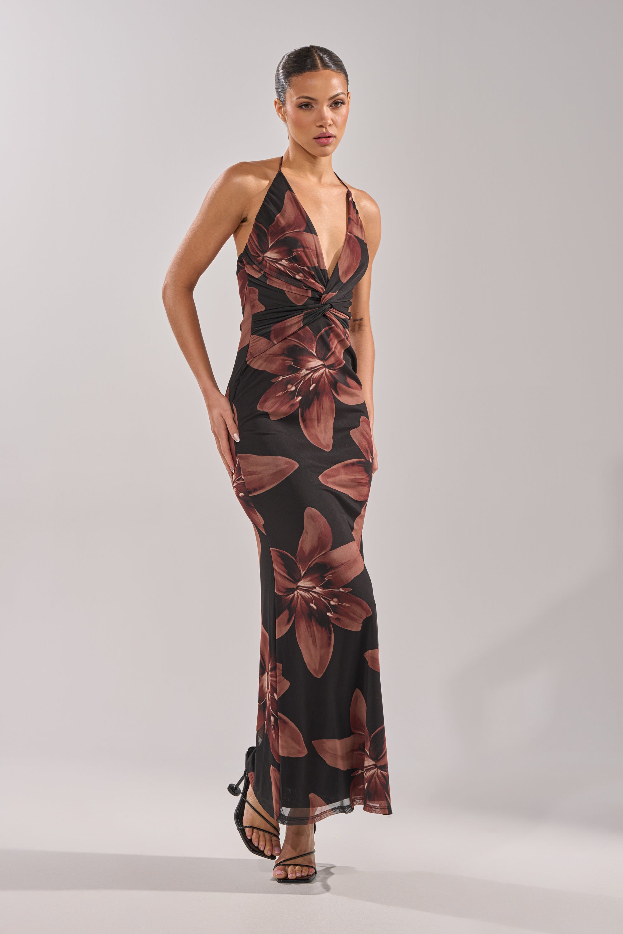 A woman confidently poses against a plain light gray background wearing the FLOWER CHILD MAXI DRESS, a sleeveless, floor-length black dress adorned with large dark red floral prints. 3