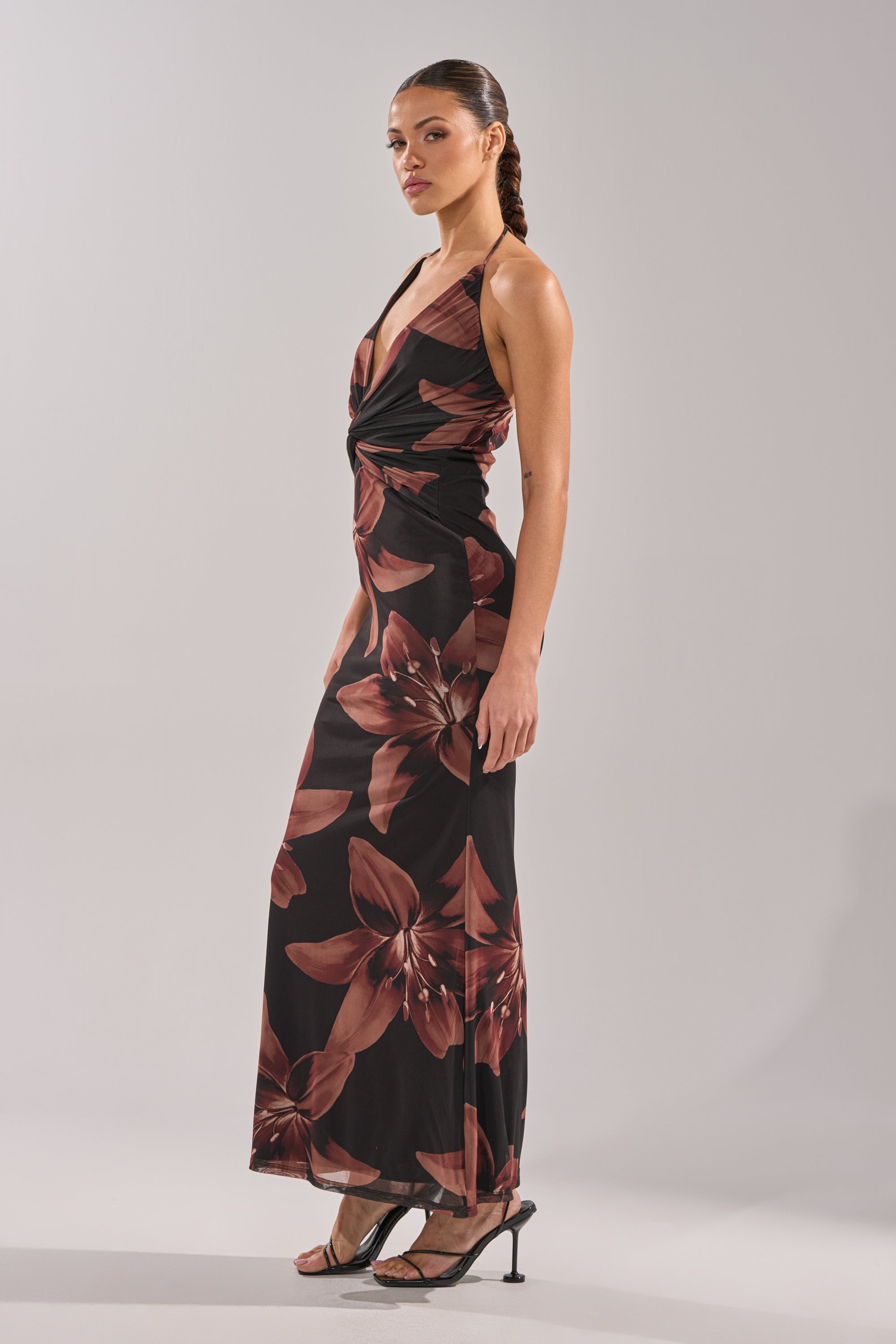 A woman wearing the FLOWER CHILD MAXI DRESS— a black sleeveless halter-neck maxi featuring large brown floral prints— stands in high-heeled sandals against a plain light background. 4