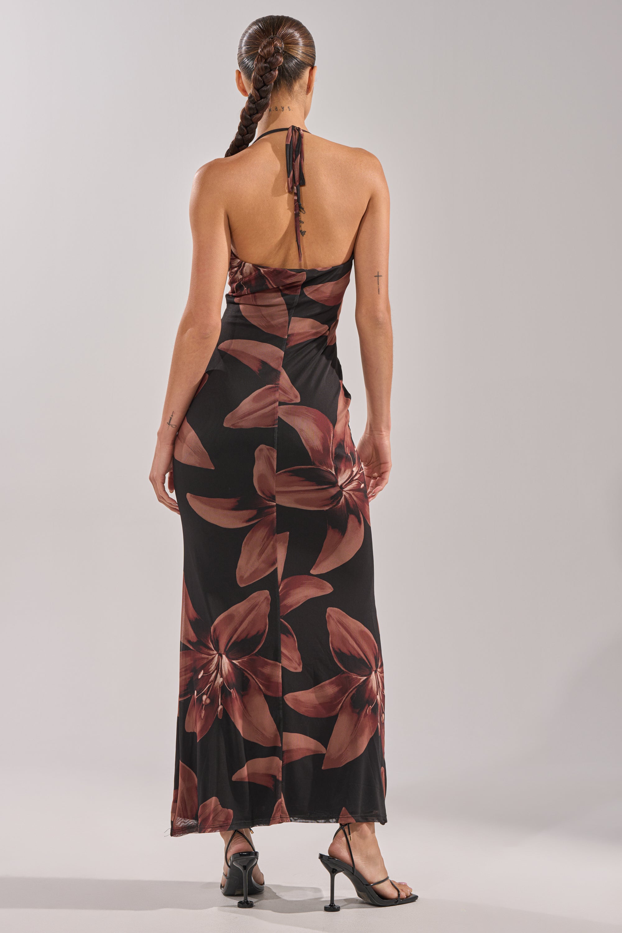 A woman with a long braid stands facing away from the camera, wearing the black, backless FLOWER CHILD MAXI DRESS with large red floral patterns and a halter neckline, paired with black heeled sandals against a plain light background. 5