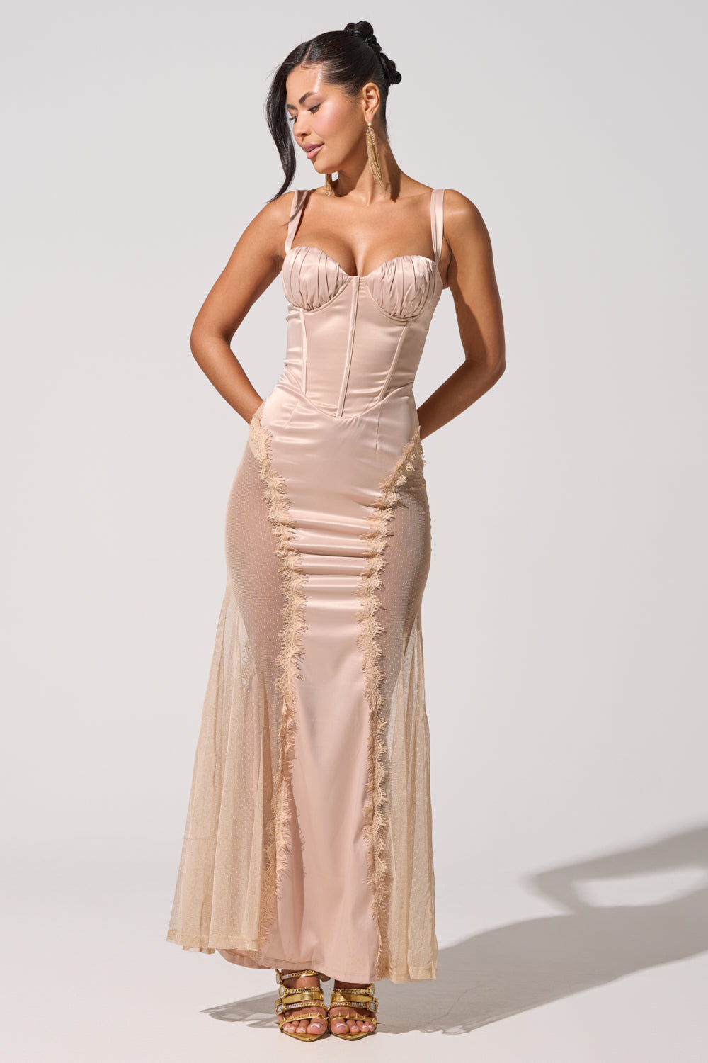 Wearing the FIND ME AT MIDNIGHT SATIN MAXI DRESS IN BLUSH, she stands elegantly in a fitted blush gown with lace mesh panels, gold heels, and long earrings. Her dark hair is in a side ponytail as she looks slightly downward. 2