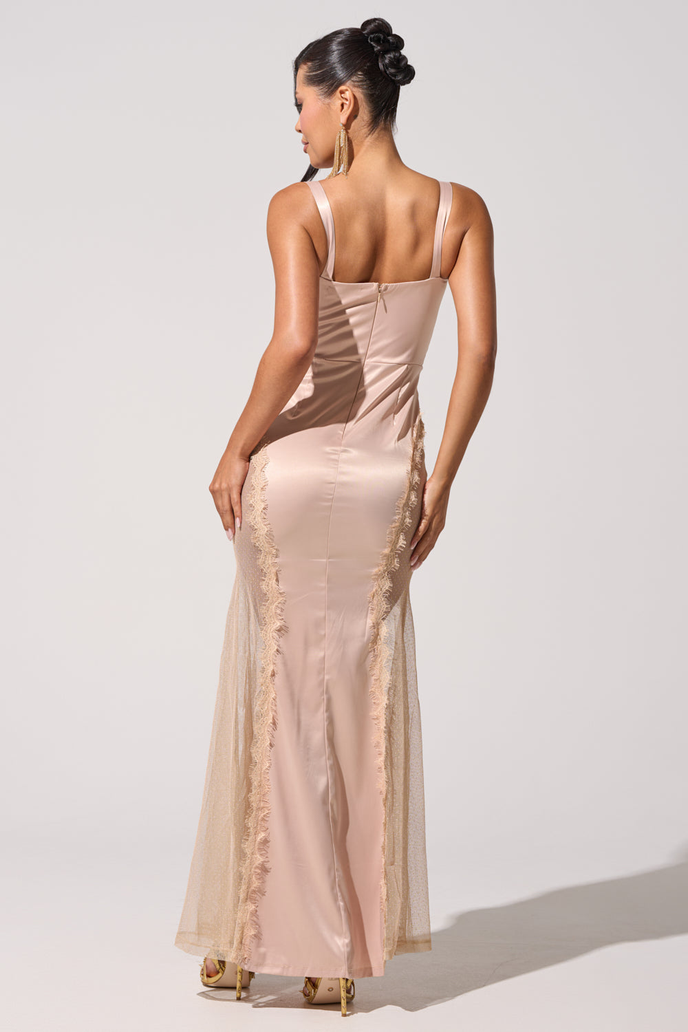 A woman stands facing away in the FIND ME AT MIDNIGHT SATIN MAXI DRESS IN BLUSH, showcasing its lace accents and elegant silhouette. Her hair is styled in an updo, complemented by gold heels and dangling earrings. 6