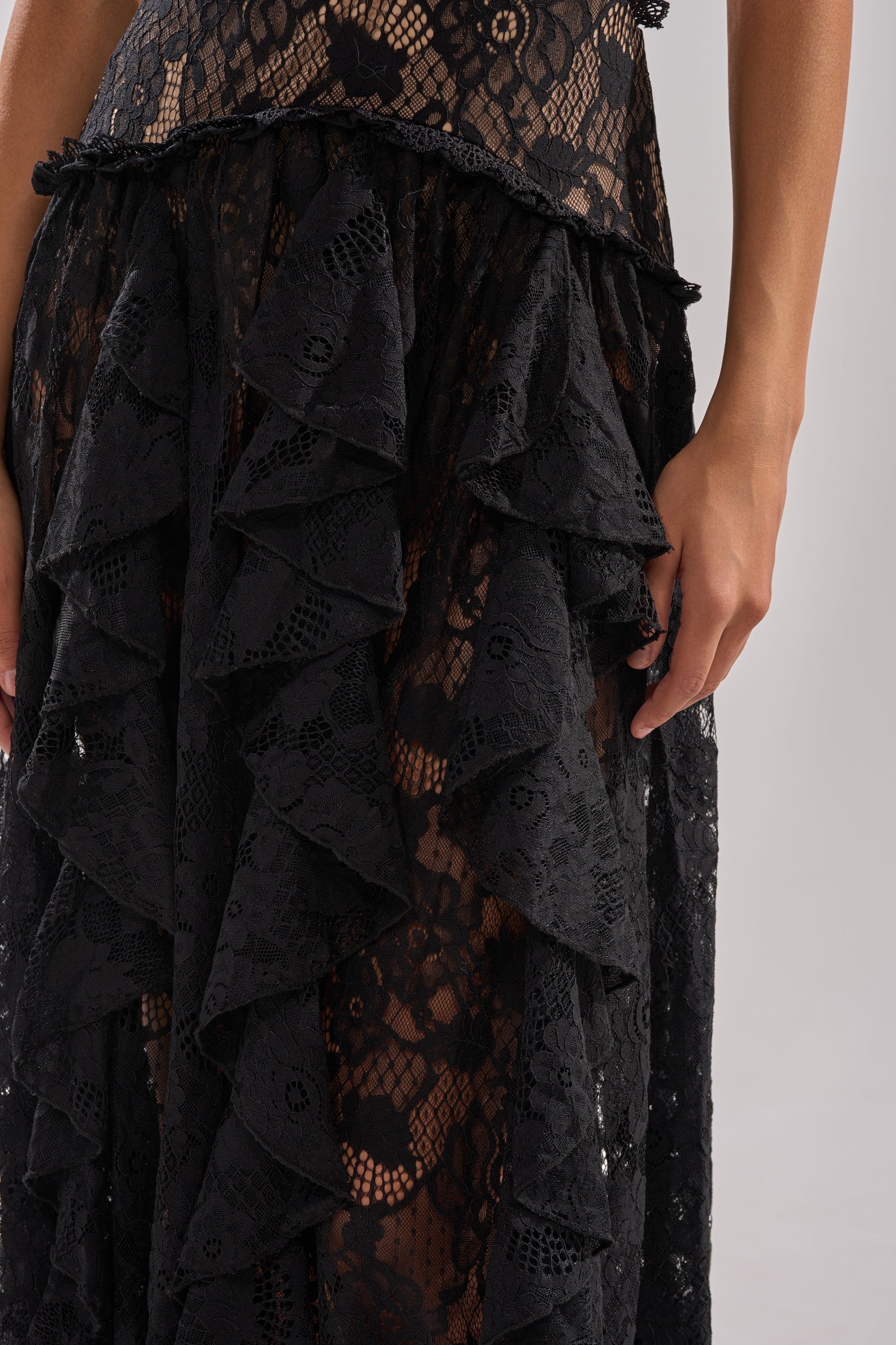 A close-up shows a person wearing the KYLA LACE MAXI DRESS IN BLACK, featuring ruffled layers and detailed floral lace. The sculpted silhouette is highlighted as their right arm rests by their side against a plain, light background.