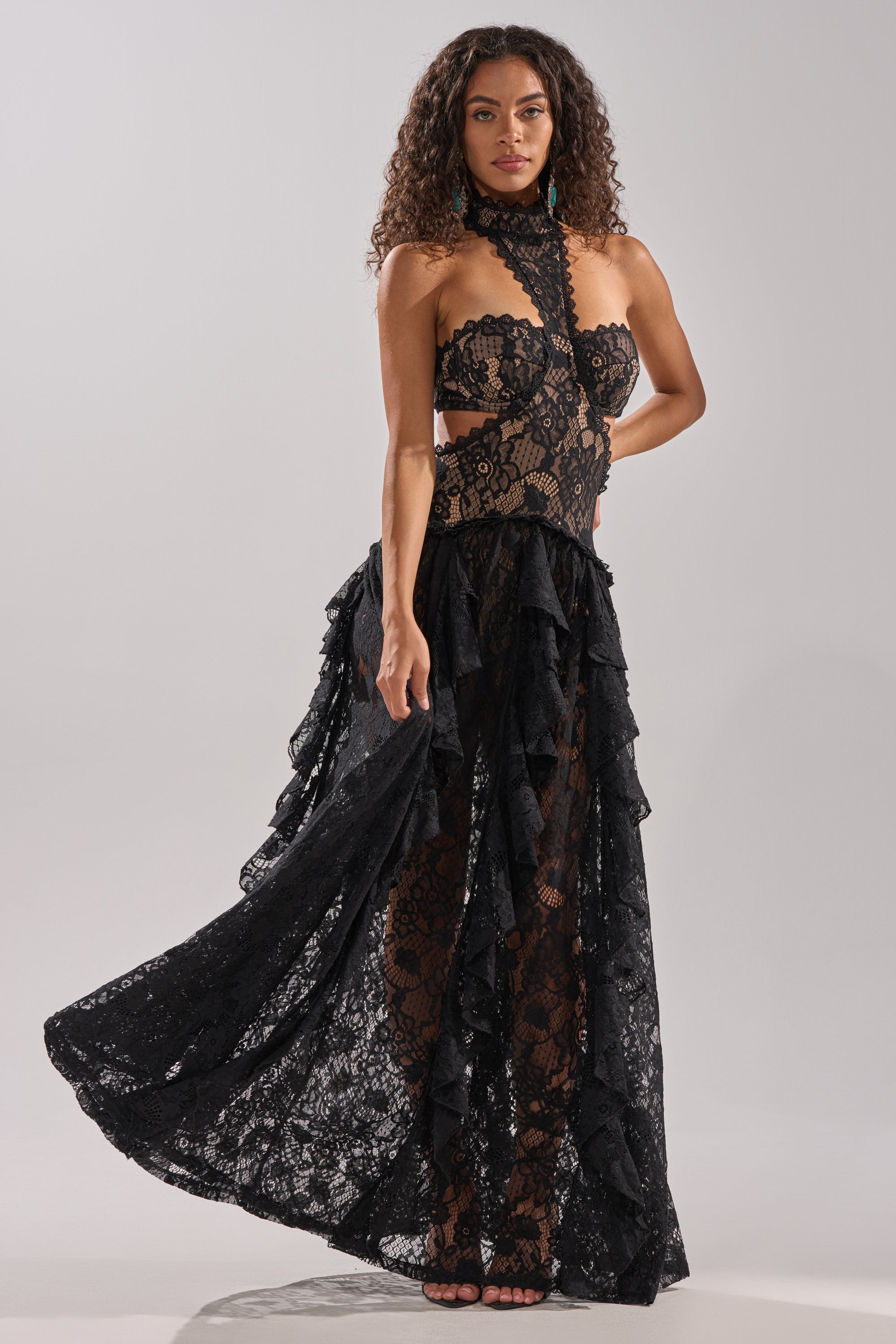 A woman with curly hair confidently models the KYLA LACE MAXI DRESS IN BLACK, a strapless gown featuring a sculpted silhouette, high neck, sheer lace panels, and ruffled details. She poses against a simple light background.