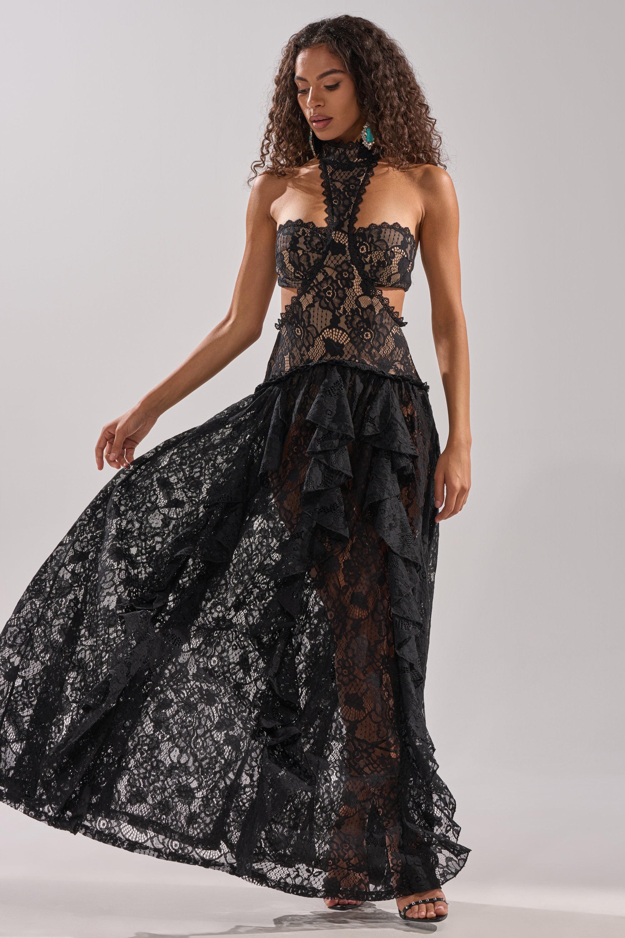 A woman poses gracefully in the KYLA LACE MAXI DRESS IN BLACK, featuring ruffle details, a high lace neckline, and a sculpted, partially sheer silhouette that subtly reveals black undergarments against a plain background.
