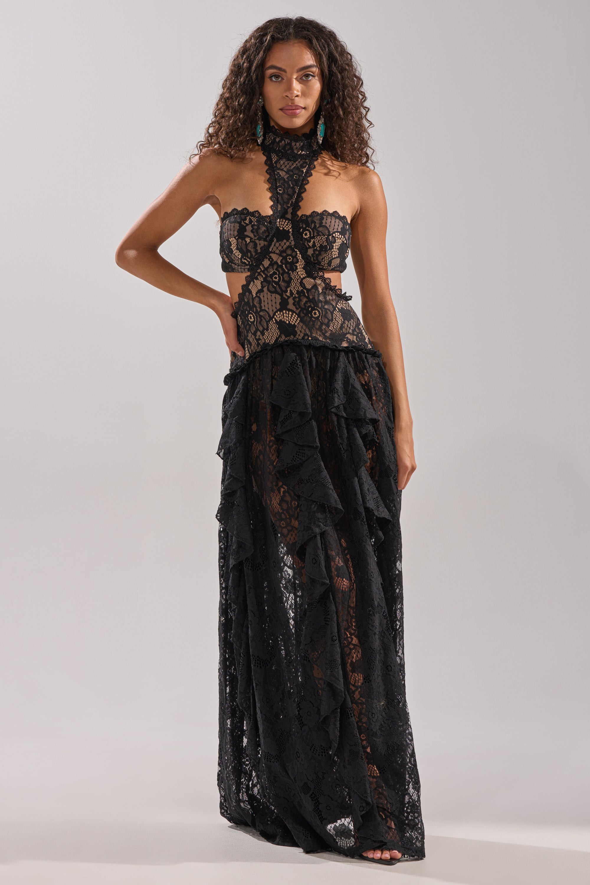 A woman models the KYLA LACE MAXI DRESS IN BLACK, showcasing its sheer panels, ruffled details, choker neckline, and sculpted silhouette. The dramatic long skirt stands out against a plain light gray background.