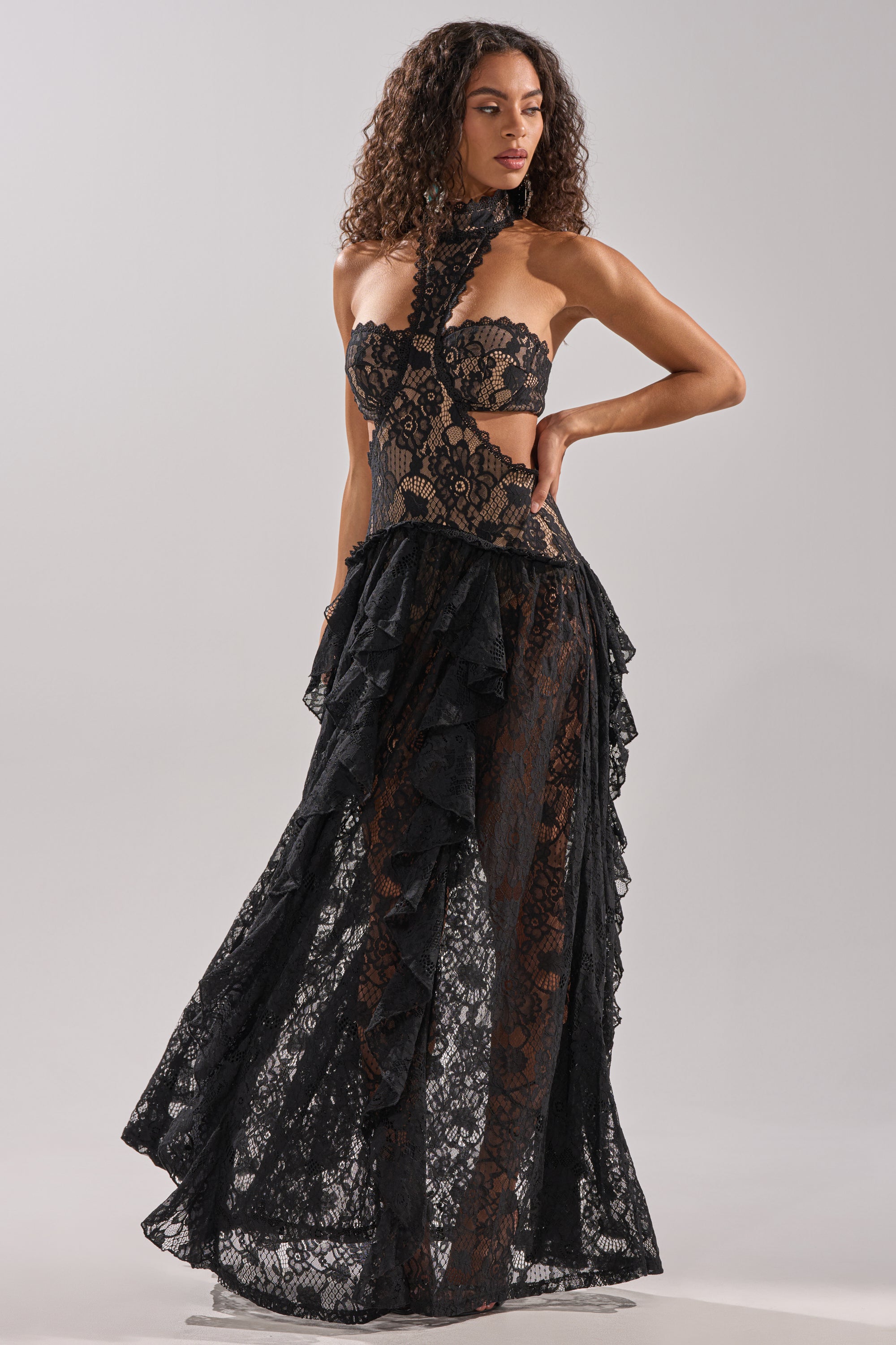 A woman wears the KYLA LACE MAXI DRESS IN BLACK, featuring cut-out sides, ruffled details, and a sheer, flowing skirt. Her poised stance and elegant silhouette stand out against a plain background as she gazes to the side.