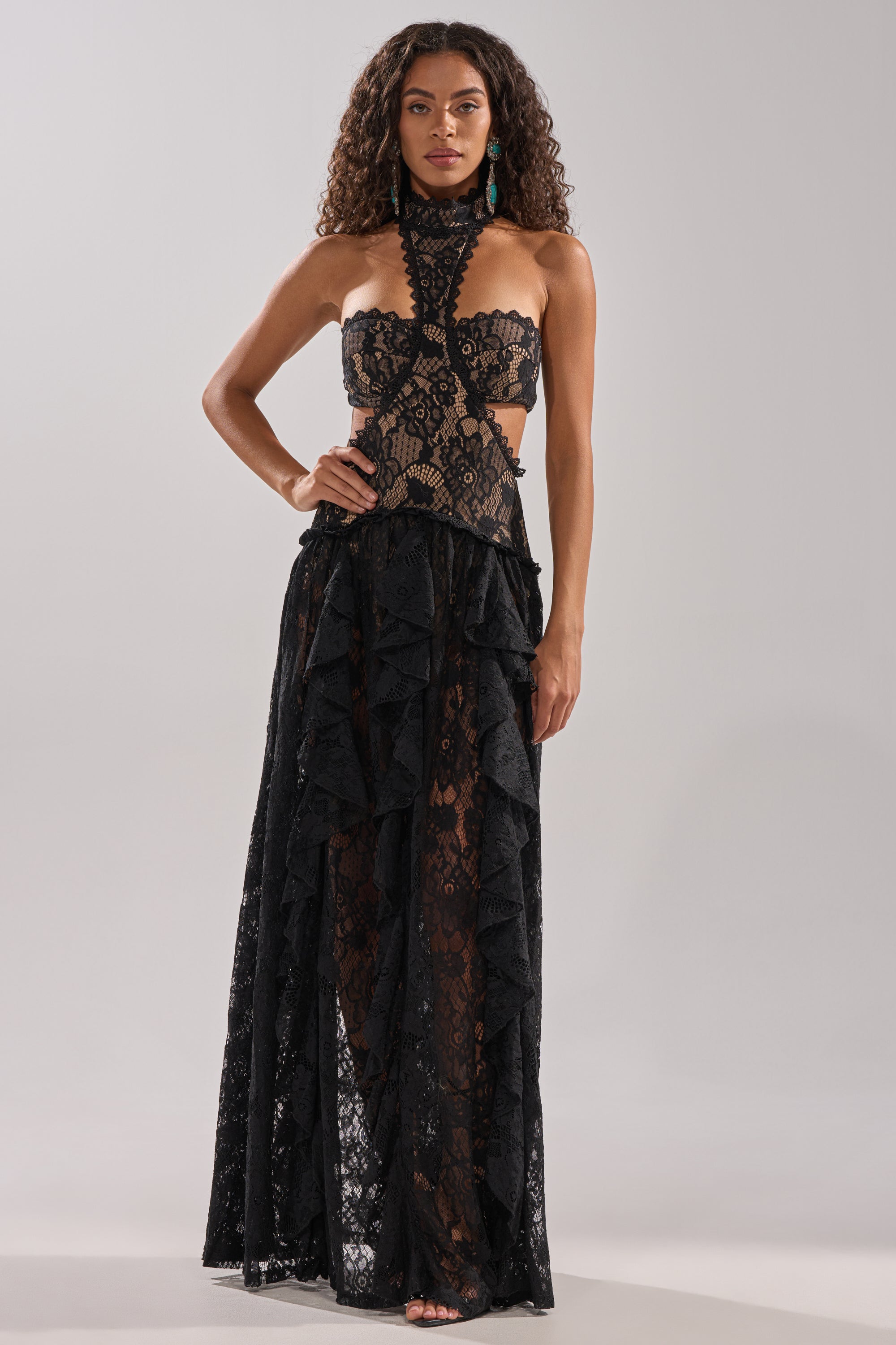 A woman with curly hair and barefoot stands facing forward against a neutral background, wearing the KYLA LACE MAXI DRESS IN BLACK featuring a sculpted silhouette, high neck, side cutouts, and a ruffled floor-length skirt.
