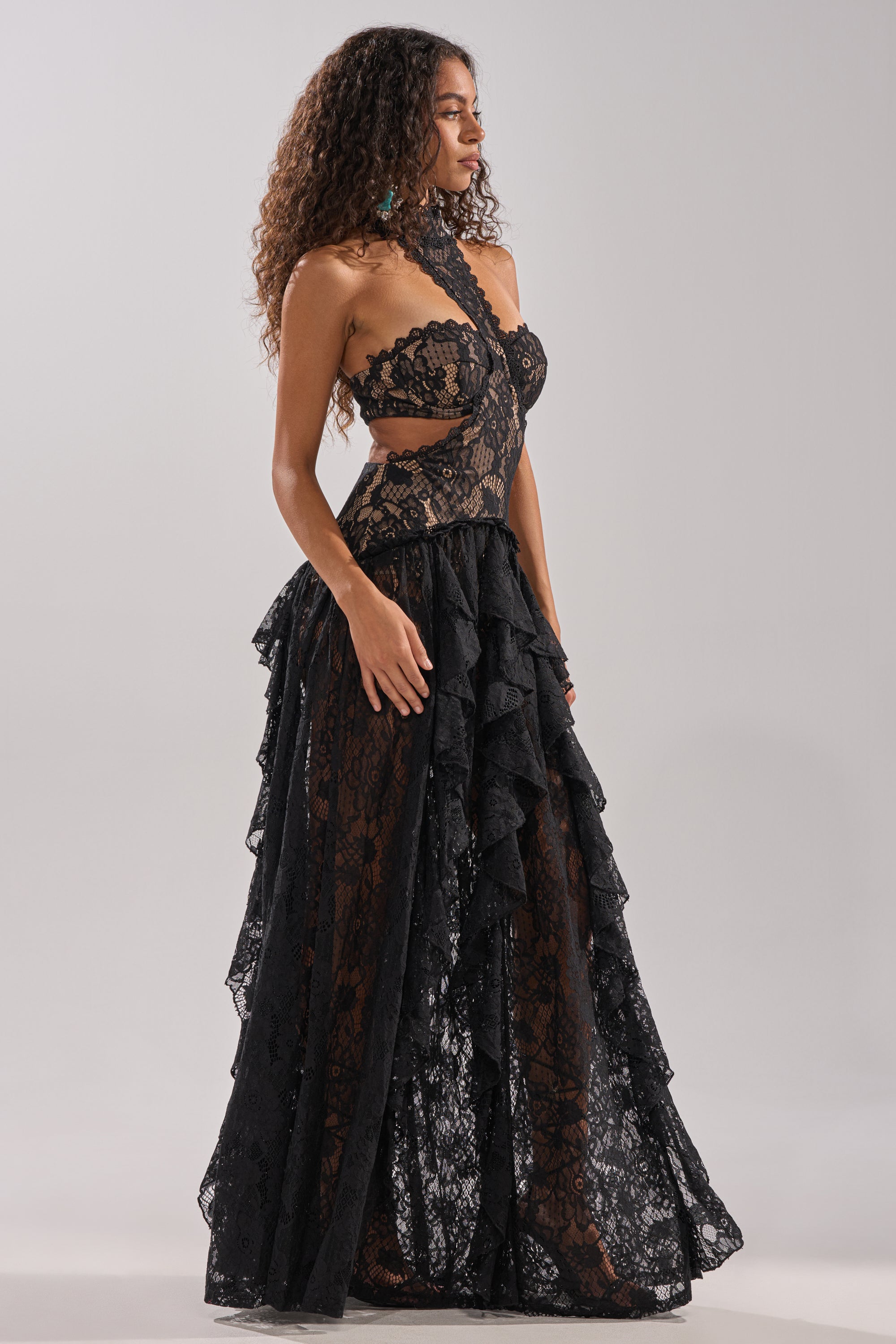 A woman with curly hair models the KYLA LACE MAXI DRESS IN BLACK— a sleeveless design featuring ruffles, cutout accents, and a sculpted fit— as she poses against a plain light gray background.