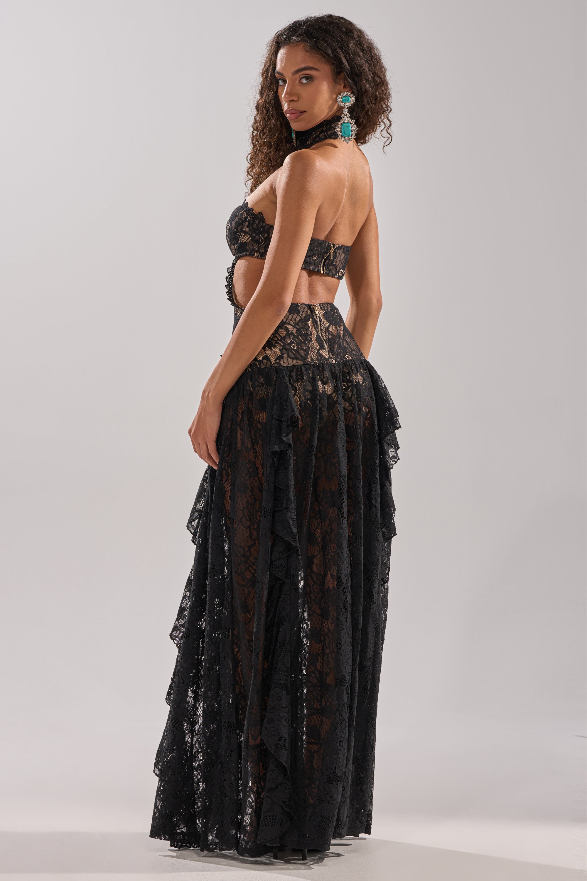 A woman with long curly hair stands sideways, looking over her shoulder in the KYLA LACE MAXI DRESS IN BLACK, a strapless gown featuring sheer panels and ruffled details. Her sculpted silhouette is accented by large turquoise earrings.