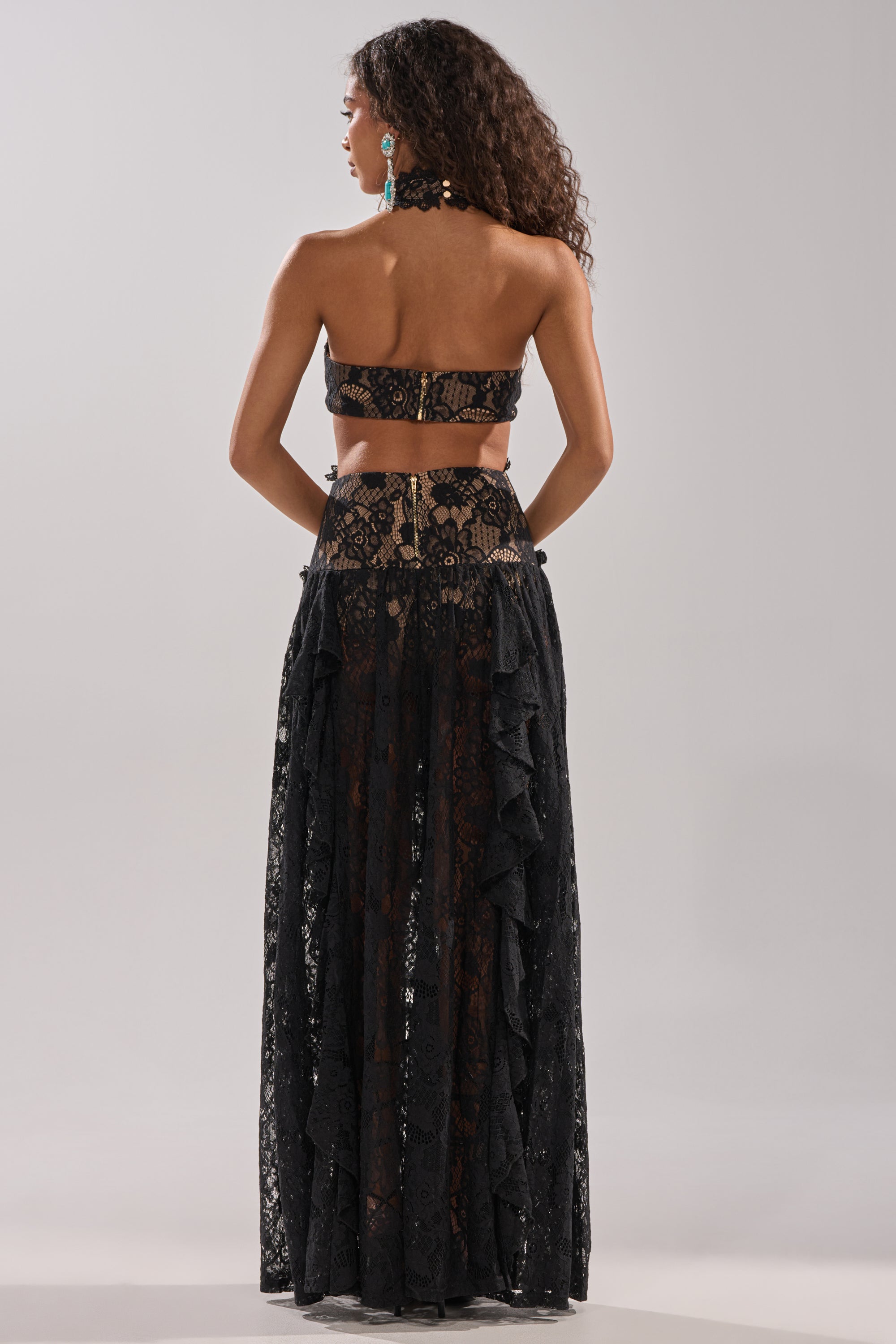 A woman with curly hair faces away, wearing the KYLA LACE MAXI DRESS IN BLACK—a halter-style lace maxi with a long, ruffled skirt that sculpts her silhouette. She pairs it with turquoise earrings and gold bracelets against a plain light gray background.