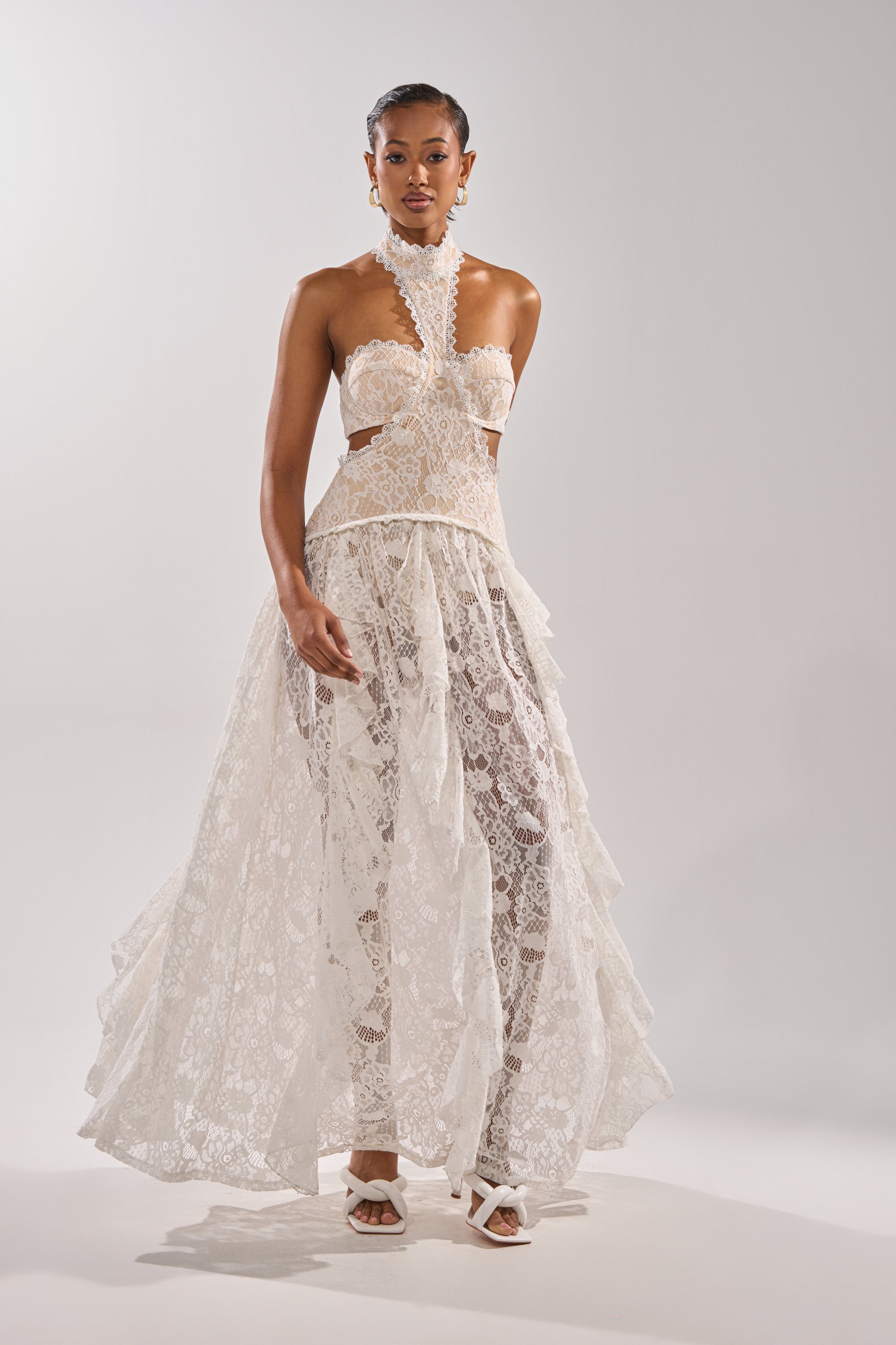 A woman models the KYLA LACE MAXI DRESS, featuring a corset-style bodice, halter neckline, and sheer layered skirt. She pairs it with white heeled sandals and earrings, her hair pulled back, posing against a plain background.