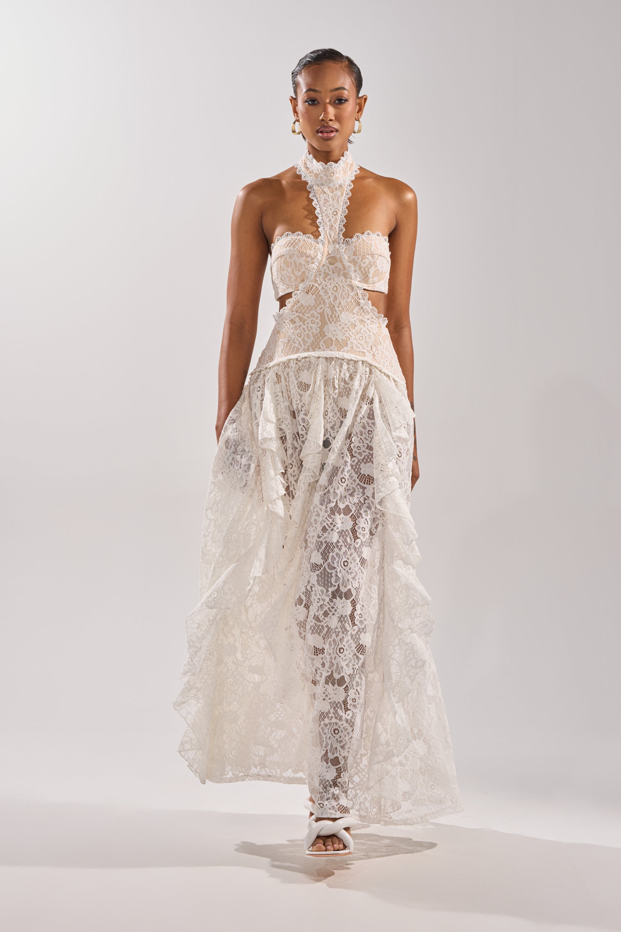 A woman stands against a plain backdrop in the KYLA LACE MAXI DRESS, featuring cut-out sides and a sheer, flowing skirt for vintage romance. She pairs it with white heels and statement earrings, gazing confidently at the camera.