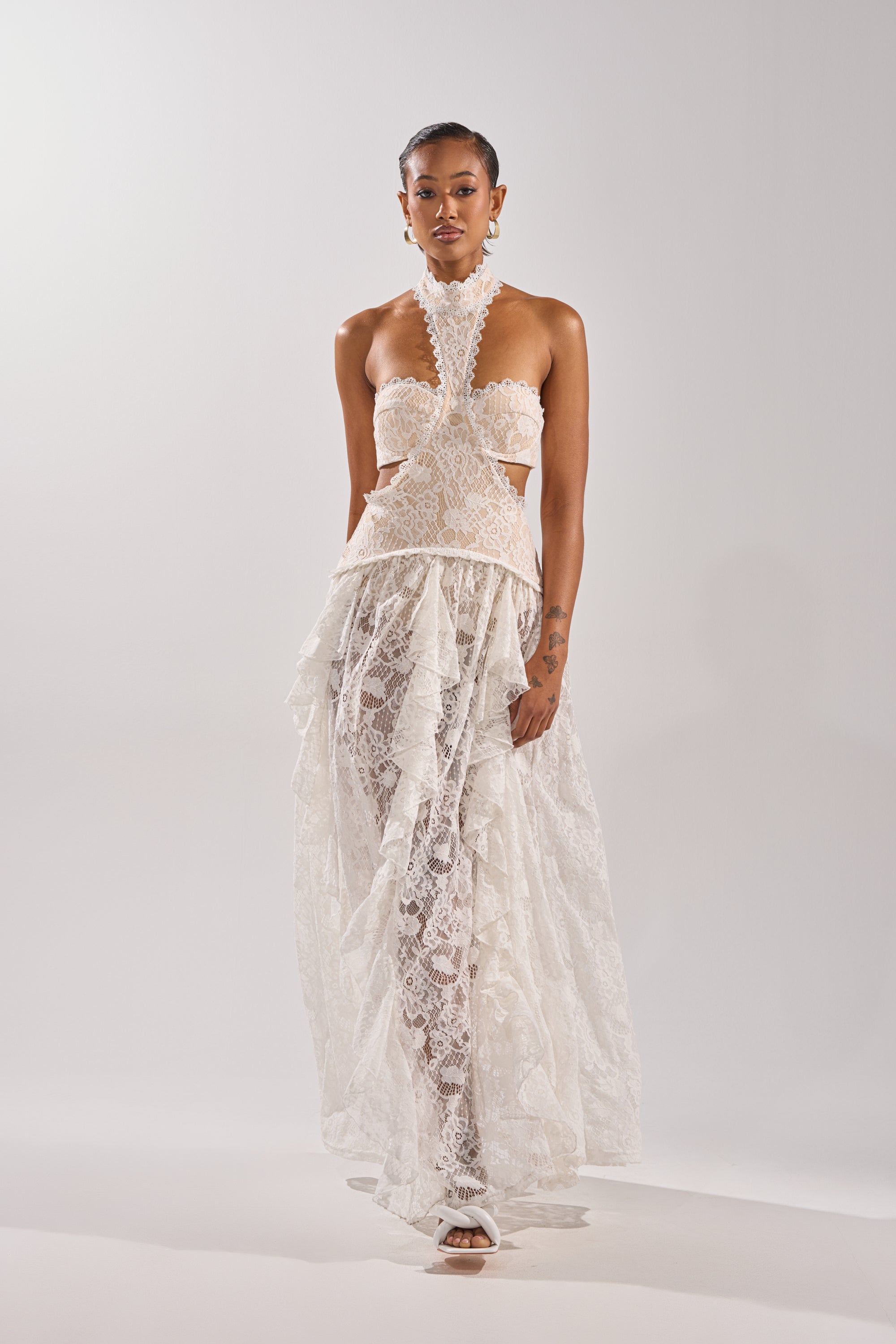 A person stands against a plain background wearing the KYLA LACE MAXI DRESS, an elegant white lace maxi with cut-out details, ruffles, a vintage-inspired halter neckline, paired with white heeled sandals.