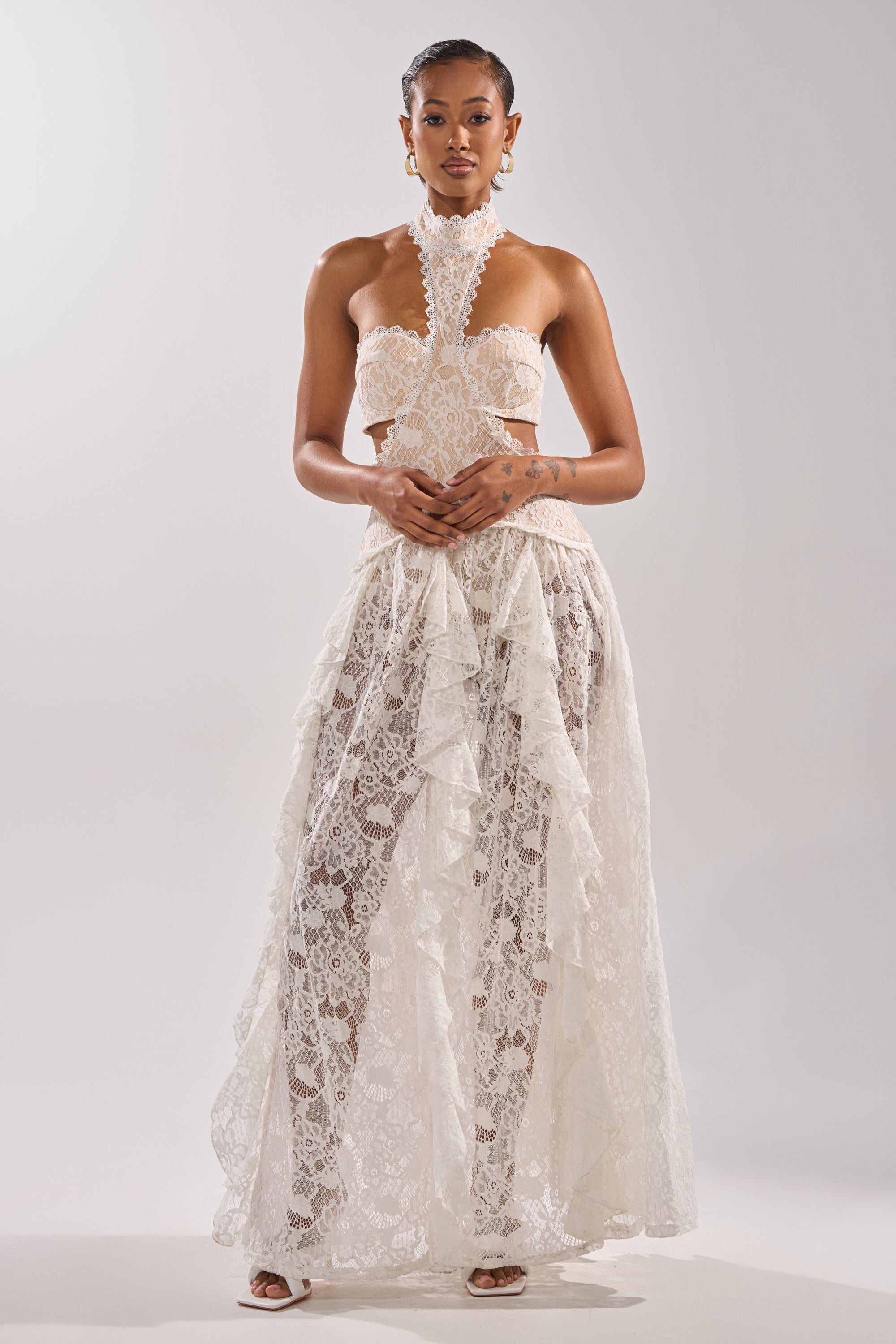 A woman with short, styled hair and statement earrings poses against a plain background in the KYLA LACE MAXI DRESS—an elegant white maxi featuring cutouts, a corset bodice, and a sheer, ruffled skirt that exudes vintage romance.