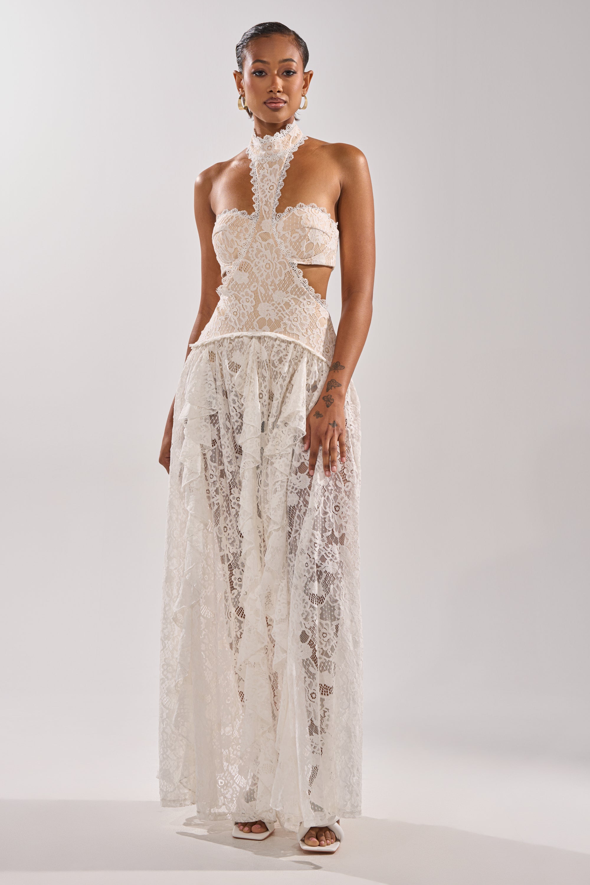 A woman poses confidently against a plain light backdrop in the KYLA LACE MAXI DRESS—sleeveless with a high neck, cut-out sides, and a sheer flowing skirt. She completes the look with white heels and slicked-back hair.