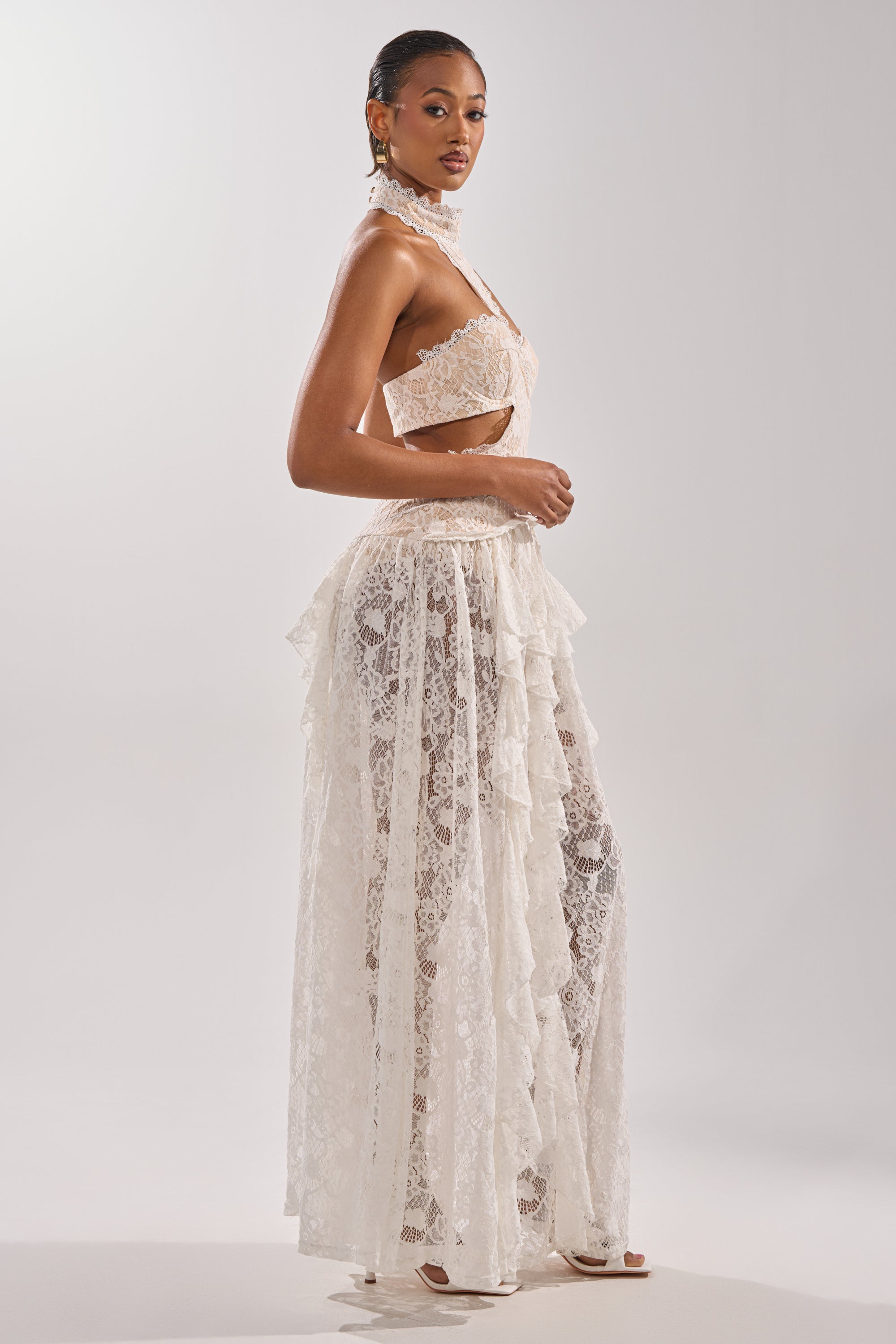 A woman poses in the KYLA LACE MAXI DRESS, a sheer white maxi gown with a halter choker top and long ruffled skirt. The plain, light background highlights this dress's elegant, intricate vintage lace details.