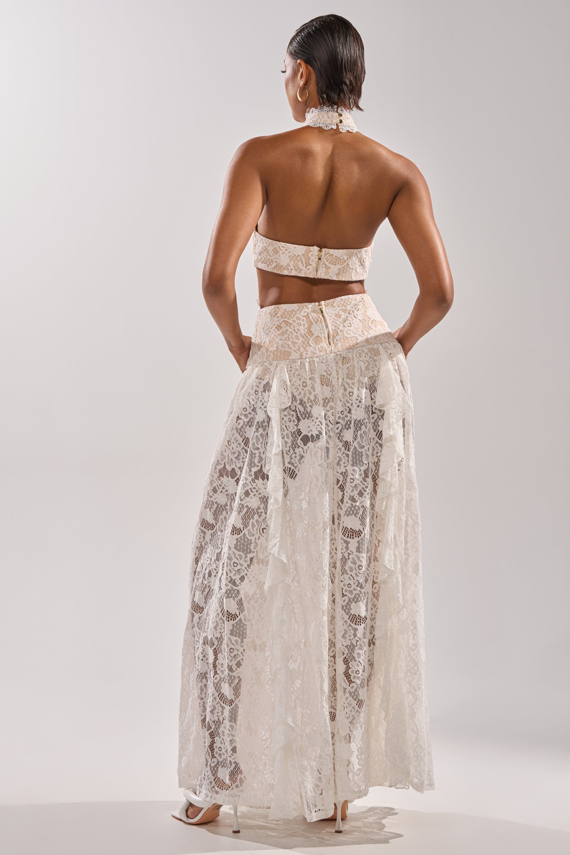 A woman faces away, wearing the KYLA LACE MAXI DRESS featuring a corset-inspired bodice and sheer details. Her hair is slicked back, and she pairs the look with white heels against a plain, light background.