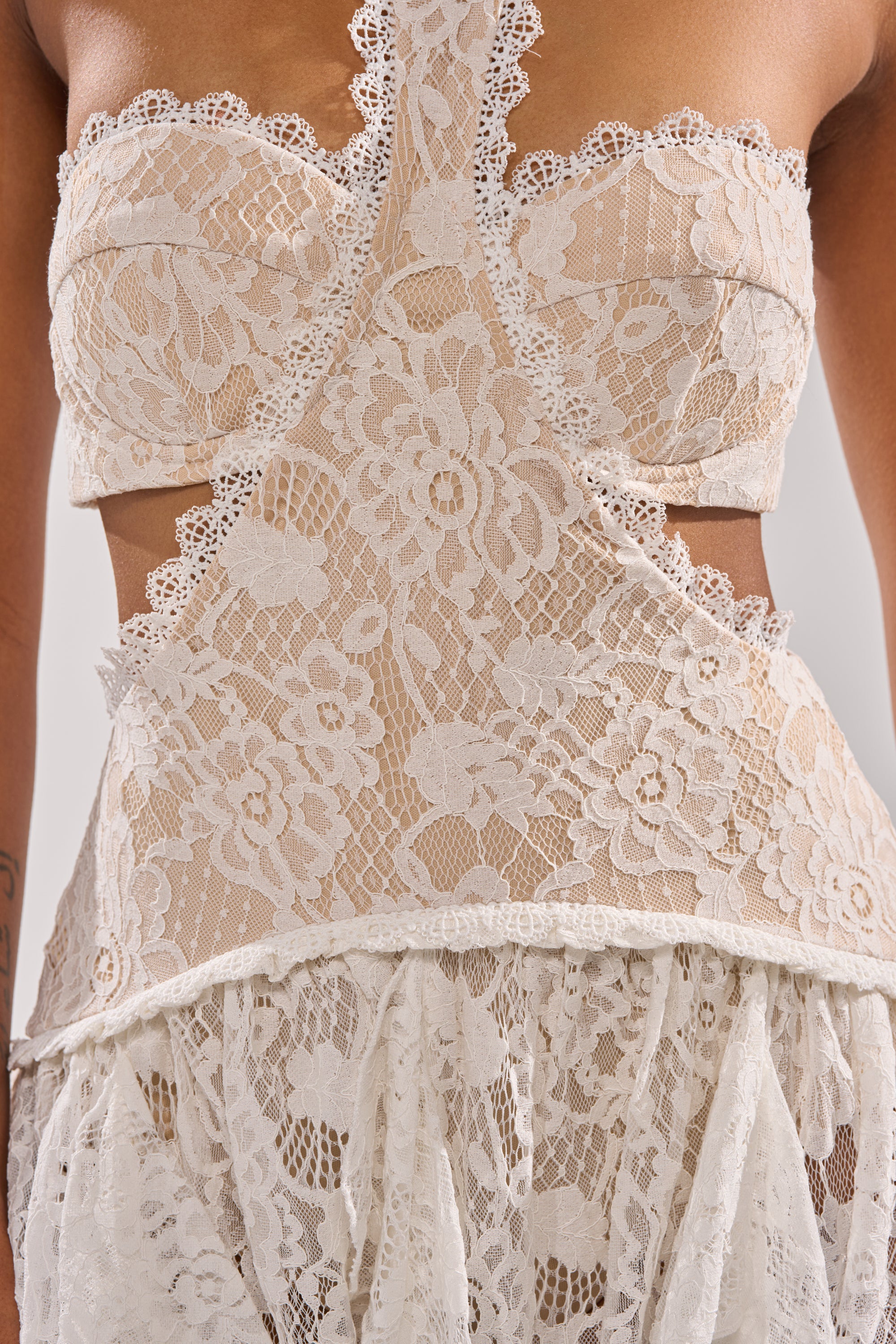 A close-up of a person wearing the KYLA LACE MAXI DRESS, featuring intricate white lace with floral patterns, cut-out waist and chest details, scalloped edges, and a corset-inspired bodice.