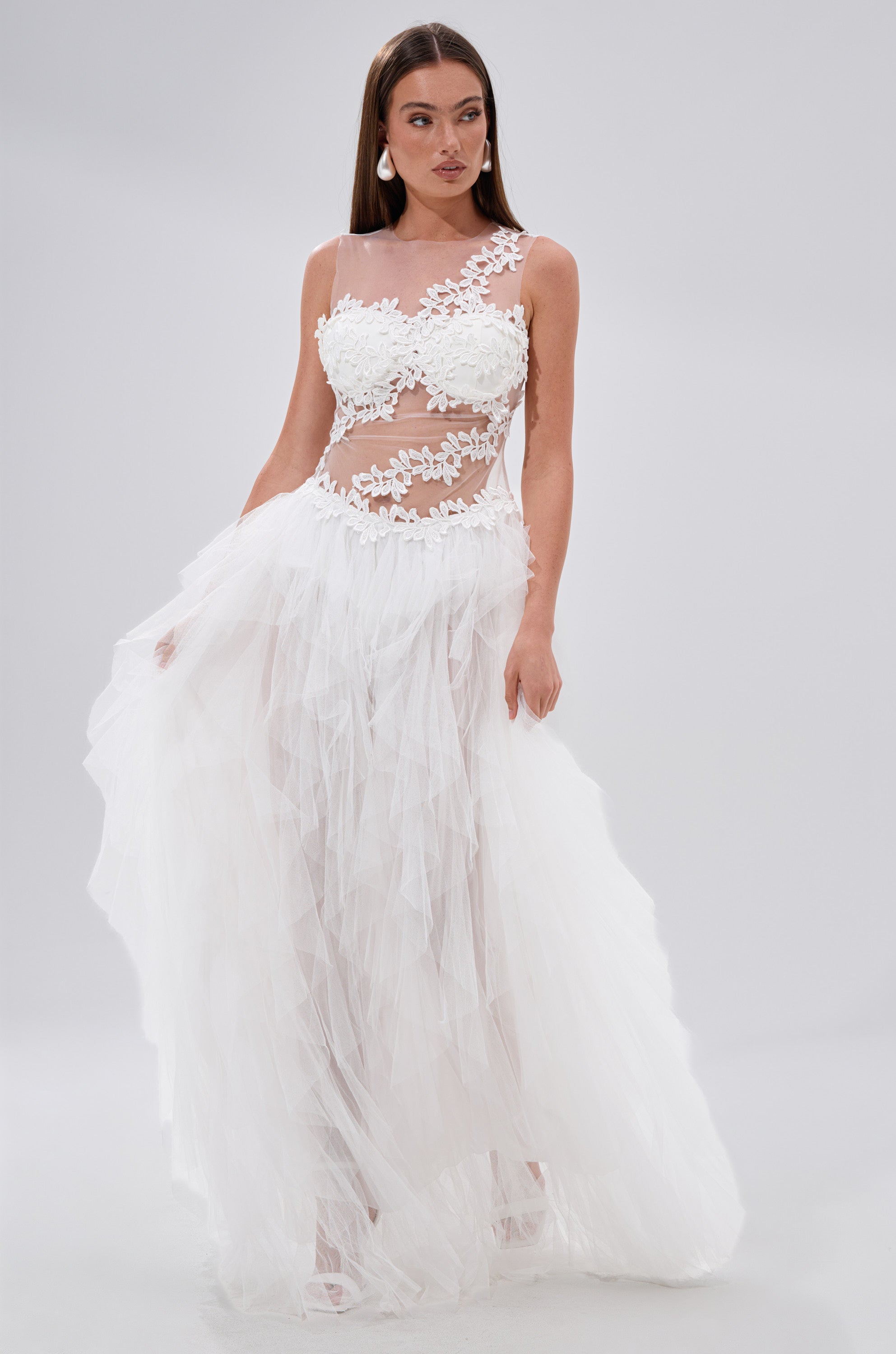 A woman stands against a light grey background wearing the FELT CUTE RUFFLE MAXI DRESS WITH FLORAL APPLIQUES, featuring a sheer mesh, floor-length ruffle design, floral appliqué bodice, one shoulder detail, and layered tulle skirt.