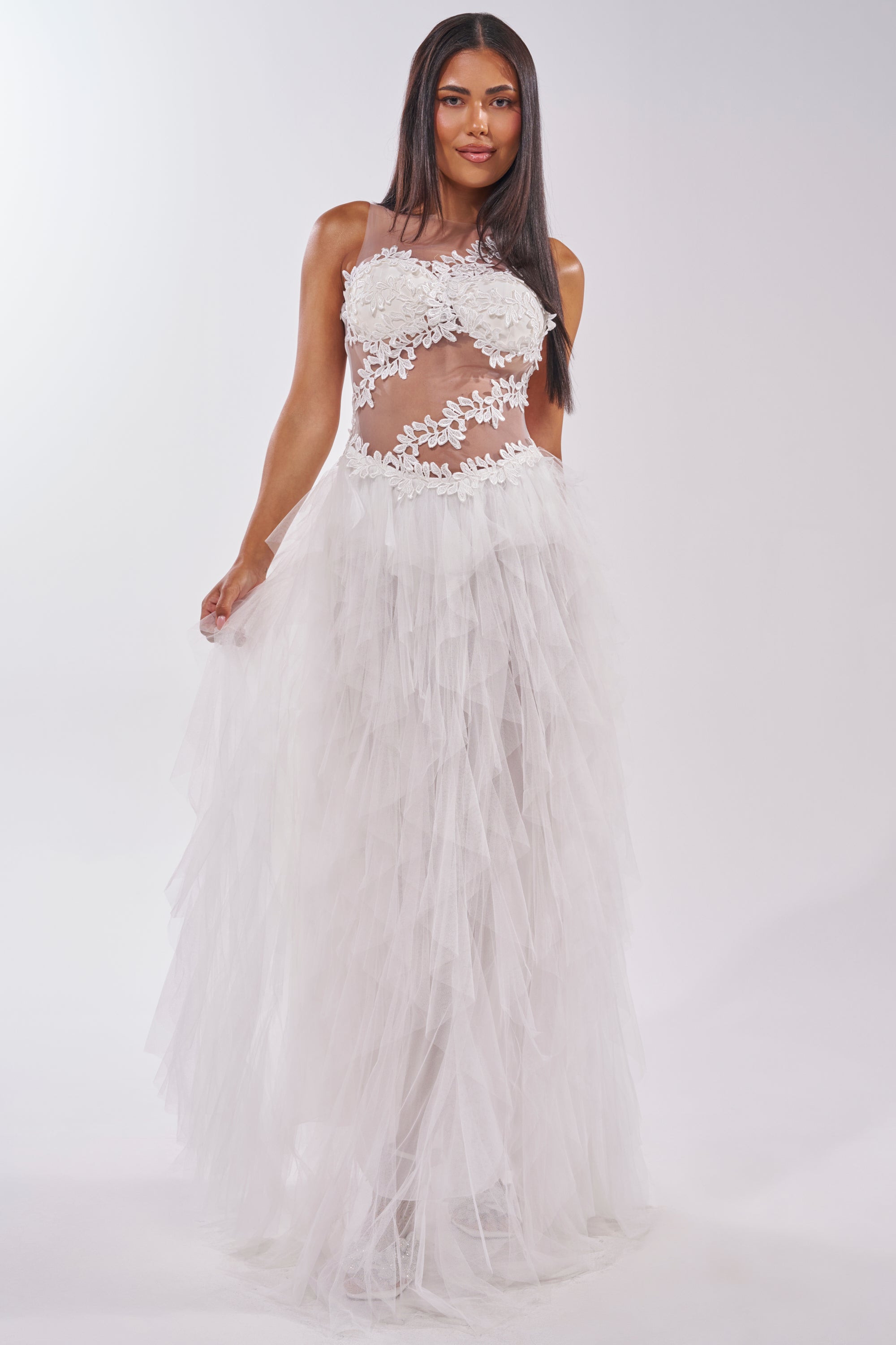 A woman poses in the FELT CUTE RUFFLE MAXI DRESS WITH FLORAL APPLIQUES, a sleeveless white gown featuring floral details on the bodice and a sheer mesh tulle skirt, creating an ethereal look against a plain light background. 0
