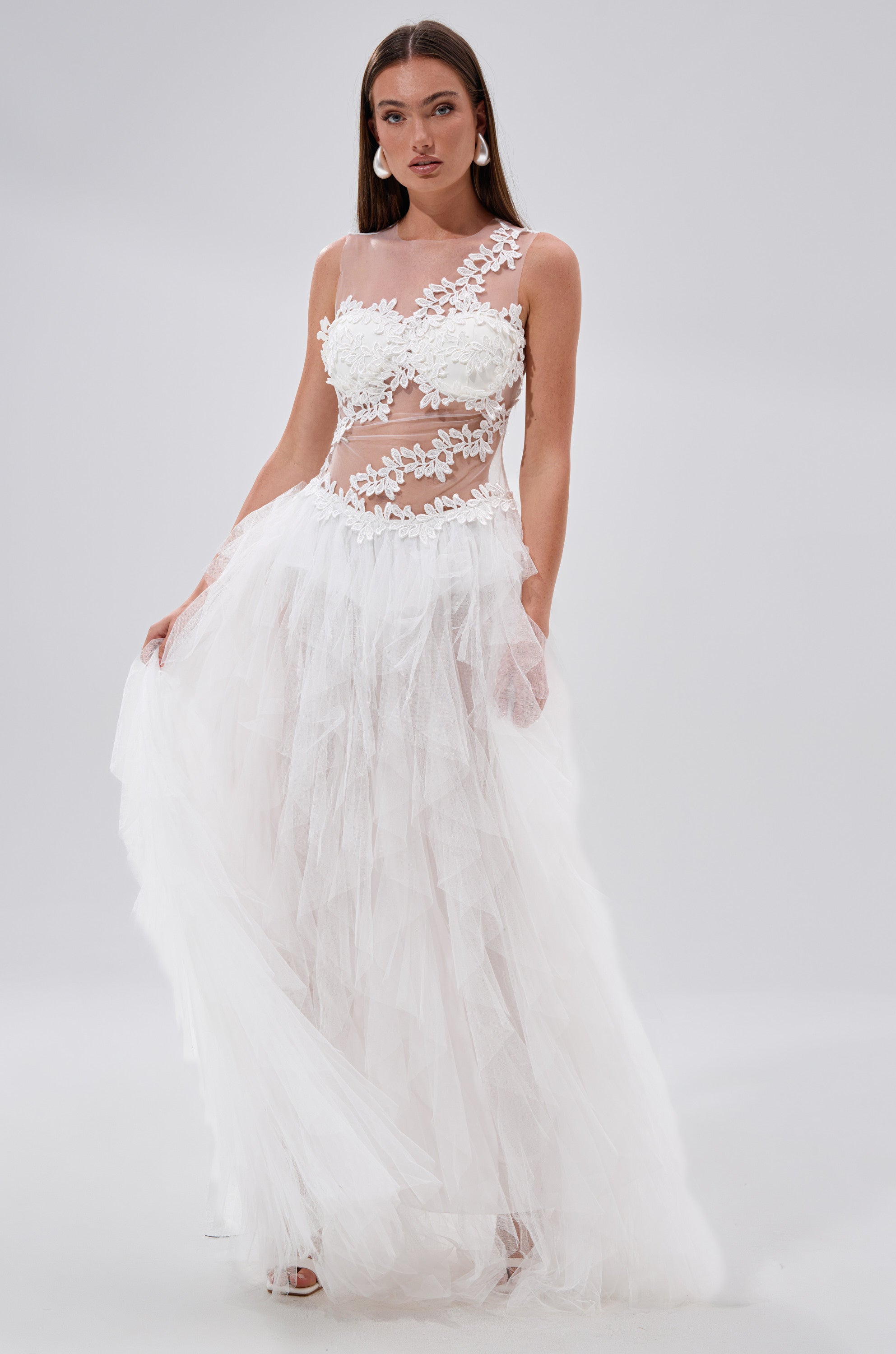 A woman poses against a plain light background in the FELT CUTE RUFFLE MAXI DRESS WITH FLORAL APPLIQUES, featuring a sheer mesh bodice adorned with floral appliques and a flowing tulle skirt that highlights its ruffled, elegant maxi style.