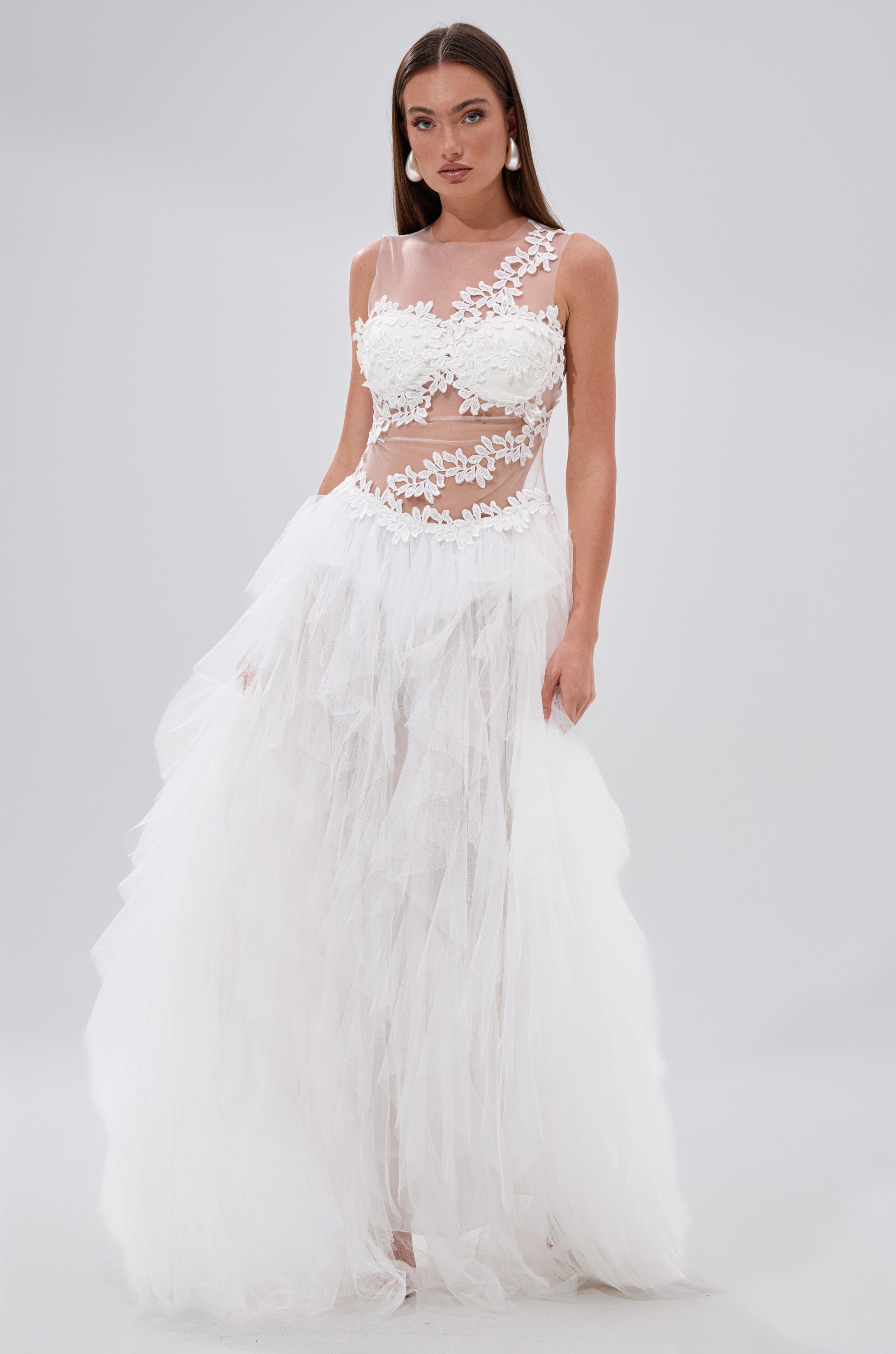 A woman stands against a plain light background wearing the FELT CUTE RUFFLE MAXI DRESS WITH FLORAL APPLIQUES, featuring a white mesh design, sheer floral-applique bodice, and a voluminous layered tulle skirt.